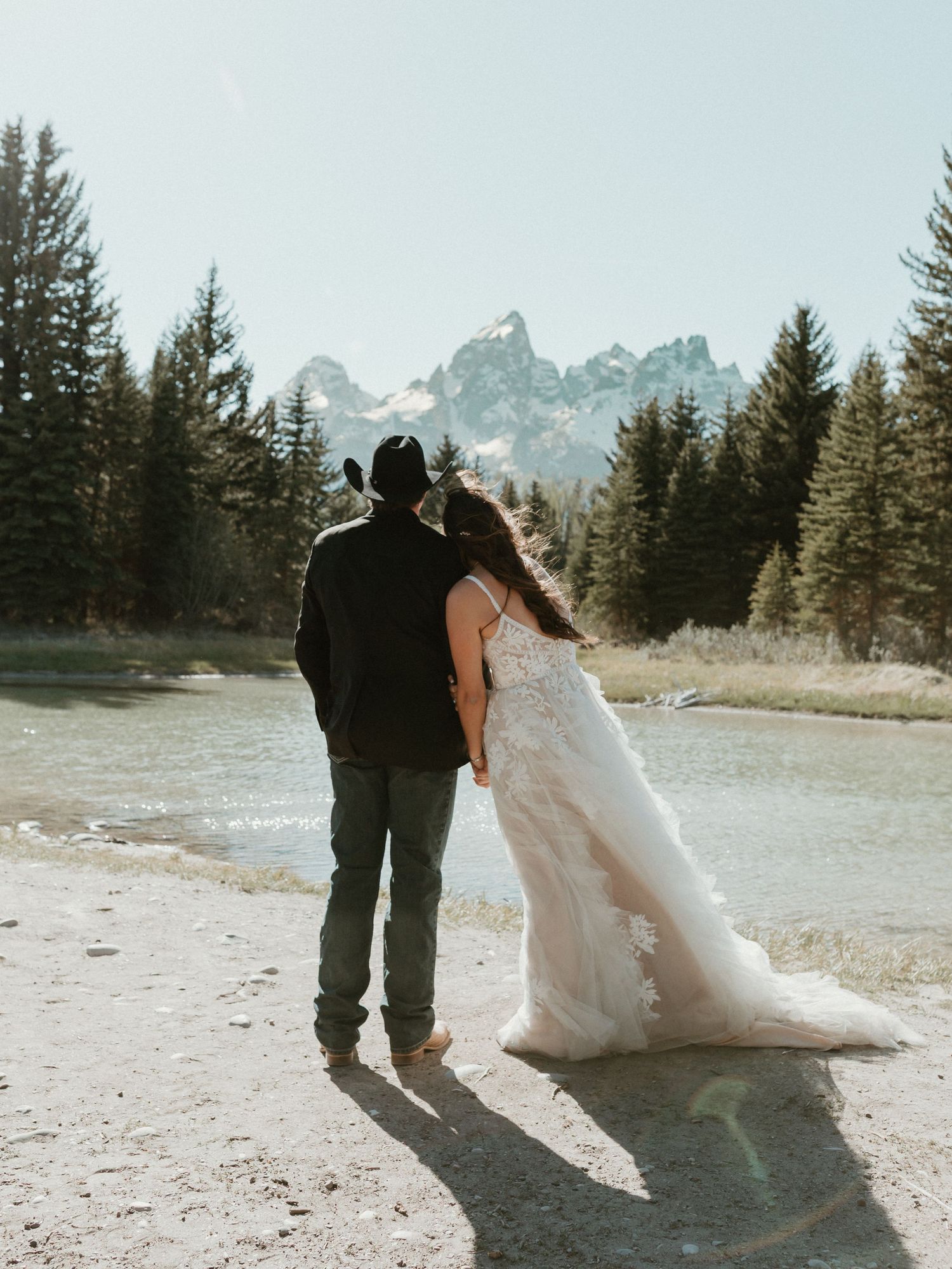 Bride in lace wedding gown and groom in cowboy hat stand by a river with Teton mountains behind them.