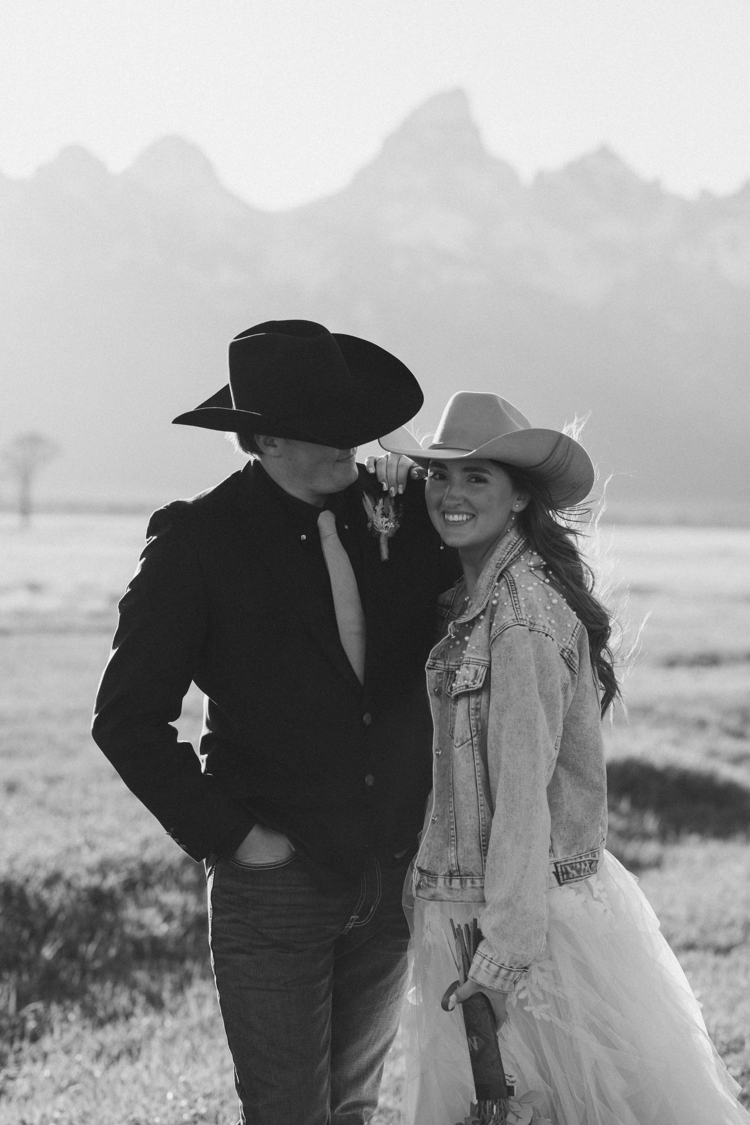 Black and white photo of couple in cowboy hats smiling together with Grand Teton mountain range behind them.