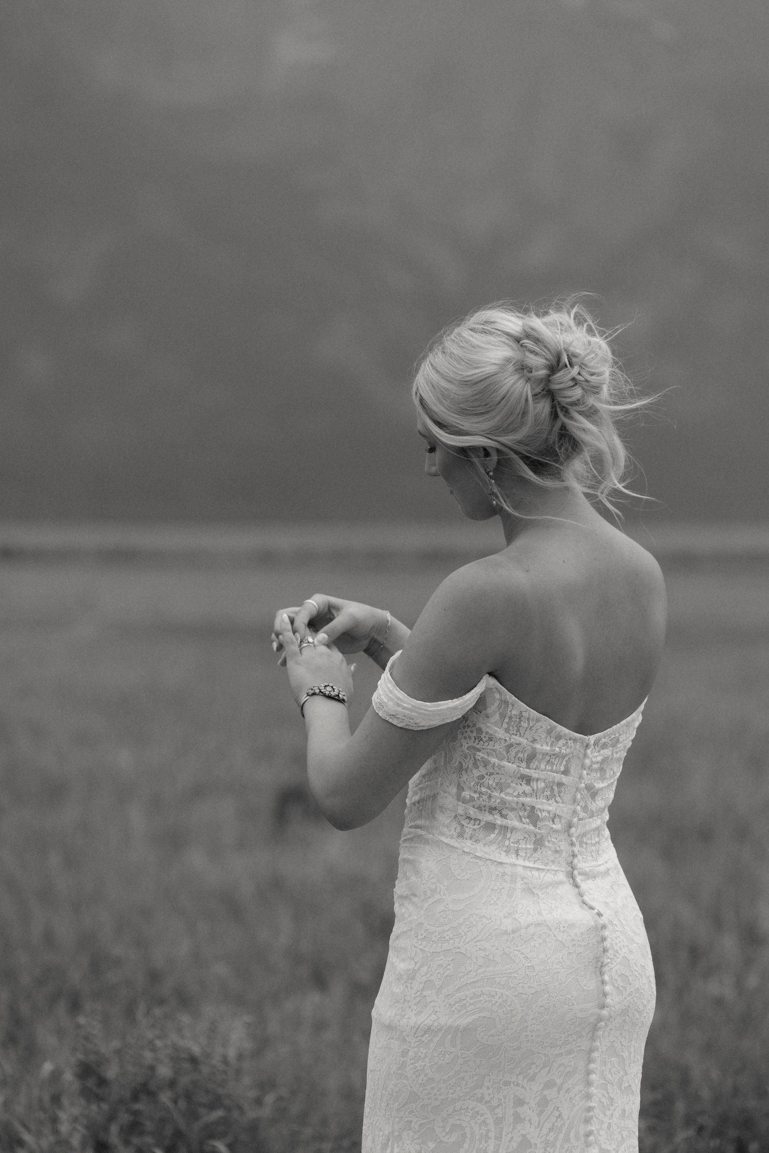 Black and white photo of a blonde bride in a lace off-shoulder gown standing in a field.