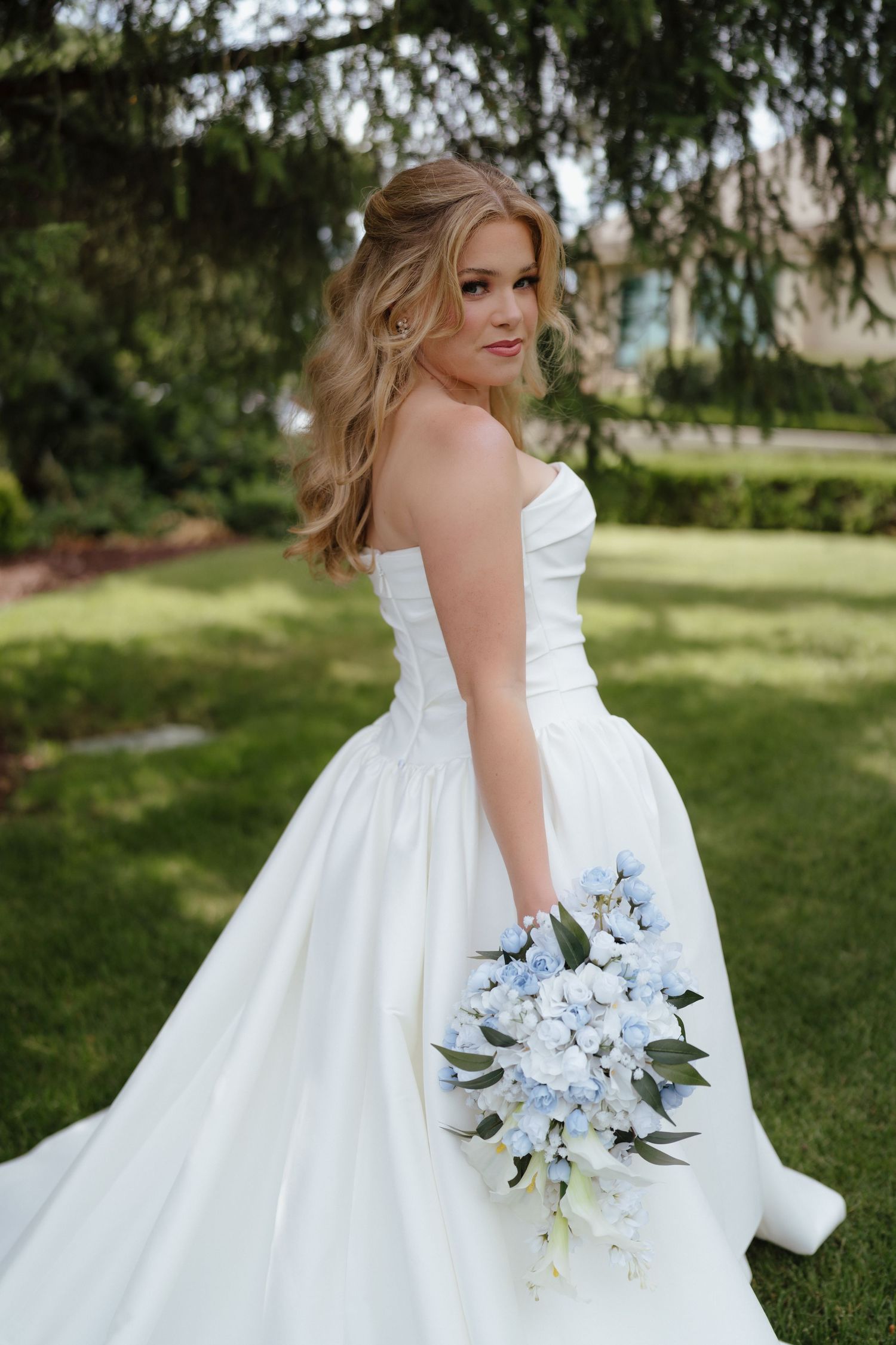 Elegant bride in a strapless white ball gown holding a cascading blue and white floral bouquet in a lush garden setting.