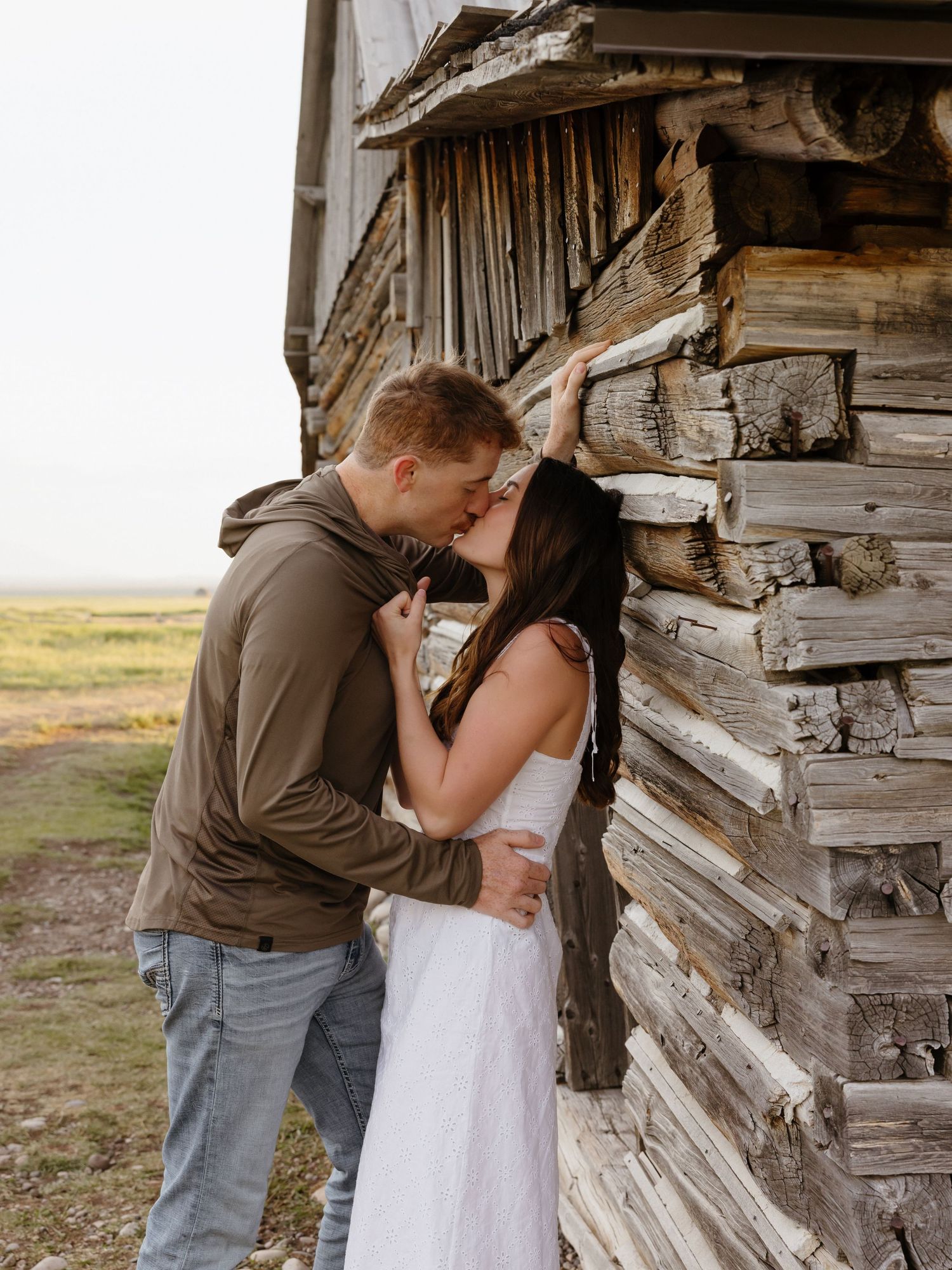 Couple sharing a kiss against a rustic weathered wooden barn wall in an open landscape.