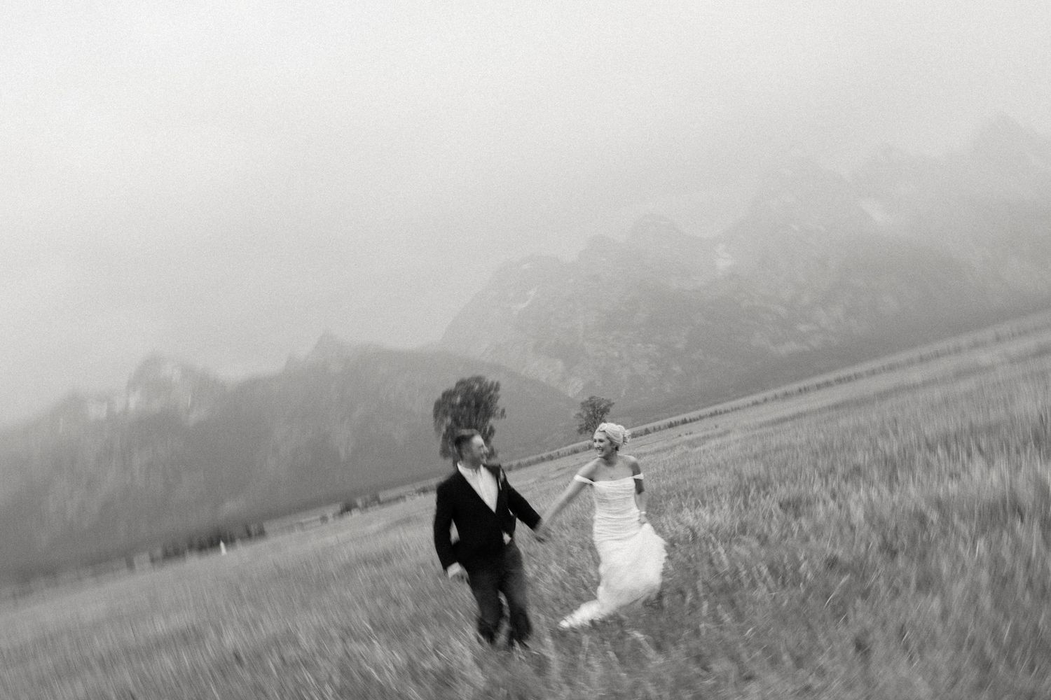 Black and white photo of a bride and groom holding hands running through a misty mountain meadow.