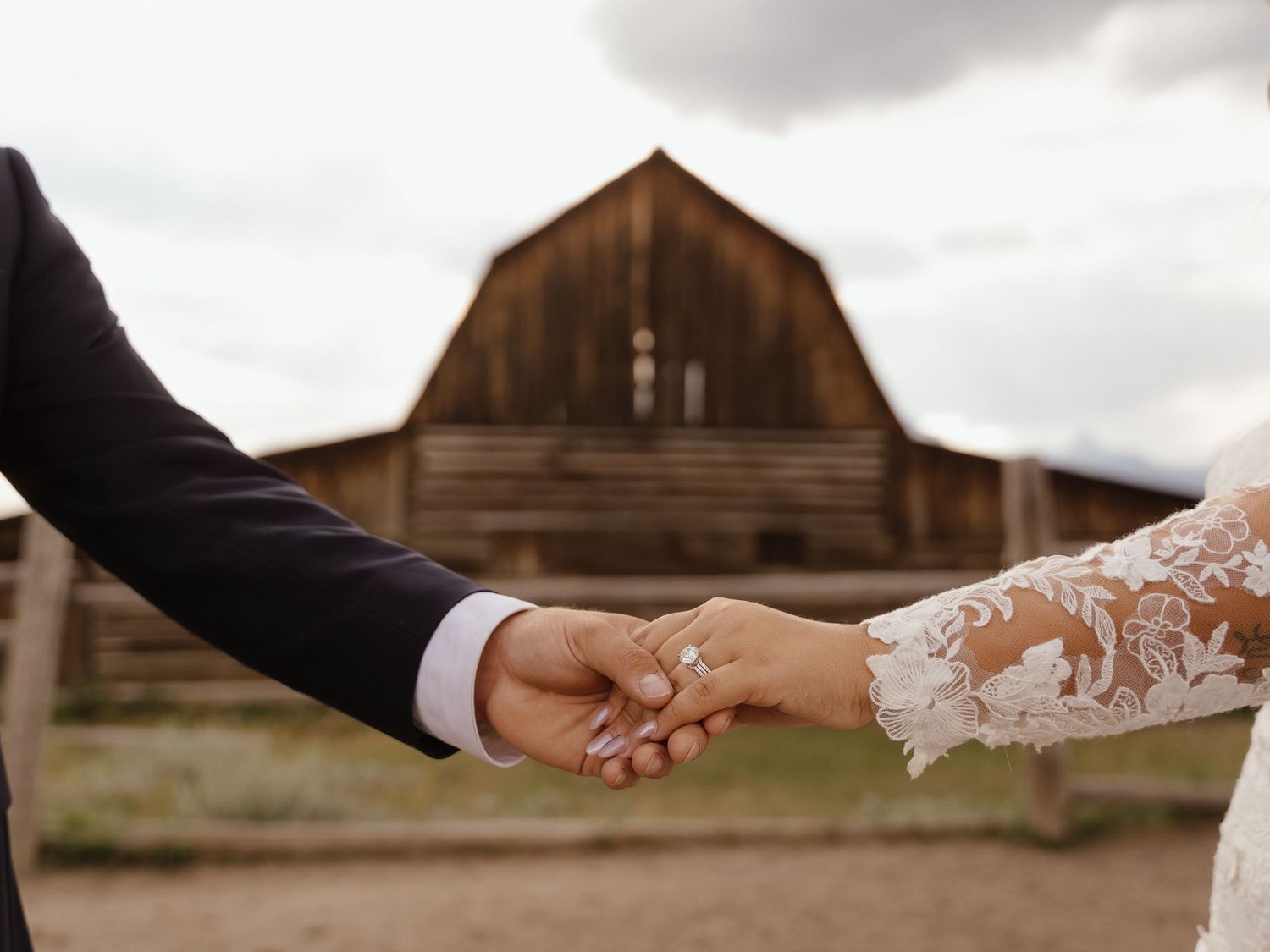 Bride and groom hold hands showing wedding ring with a rustic wooden barn in the background.