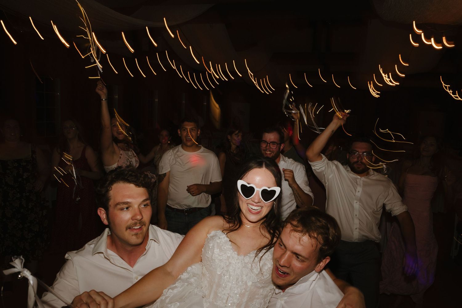 Bride in heart sunglasses celebrates with guests during lively wedding reception dance party.
