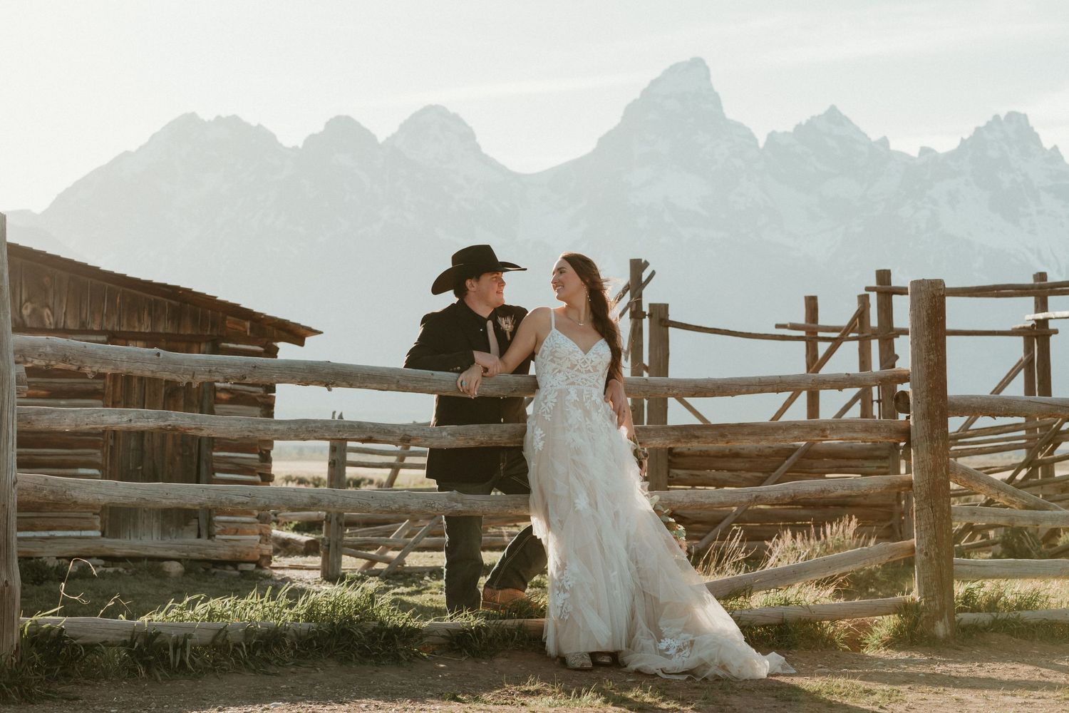 Bride and groom sharing a romantic moment at a rustic fence with Grand Teton mountains behind.