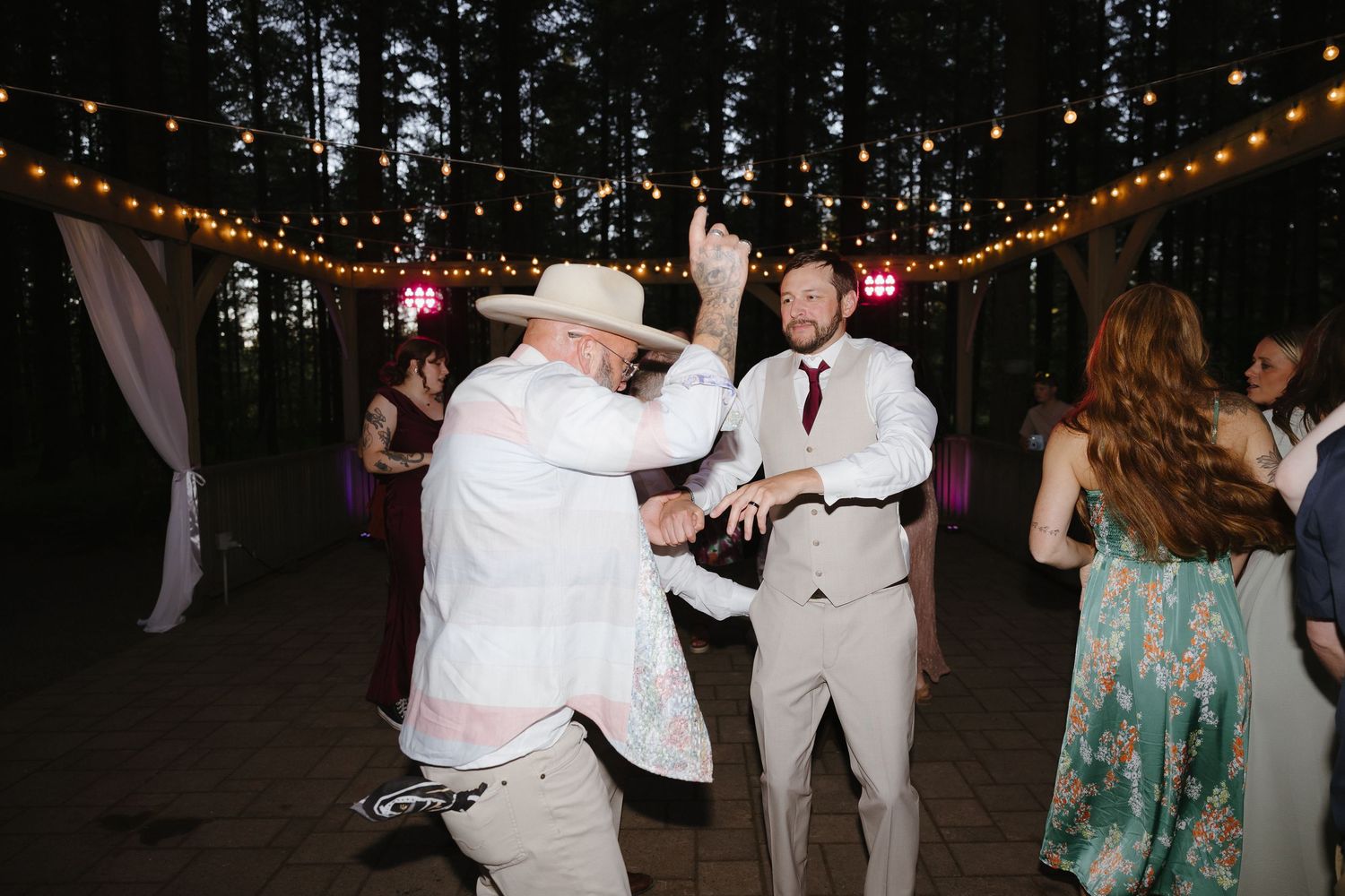 Wedding guests dancing energetically under string lights at an outdoor forest wedding reception at night.