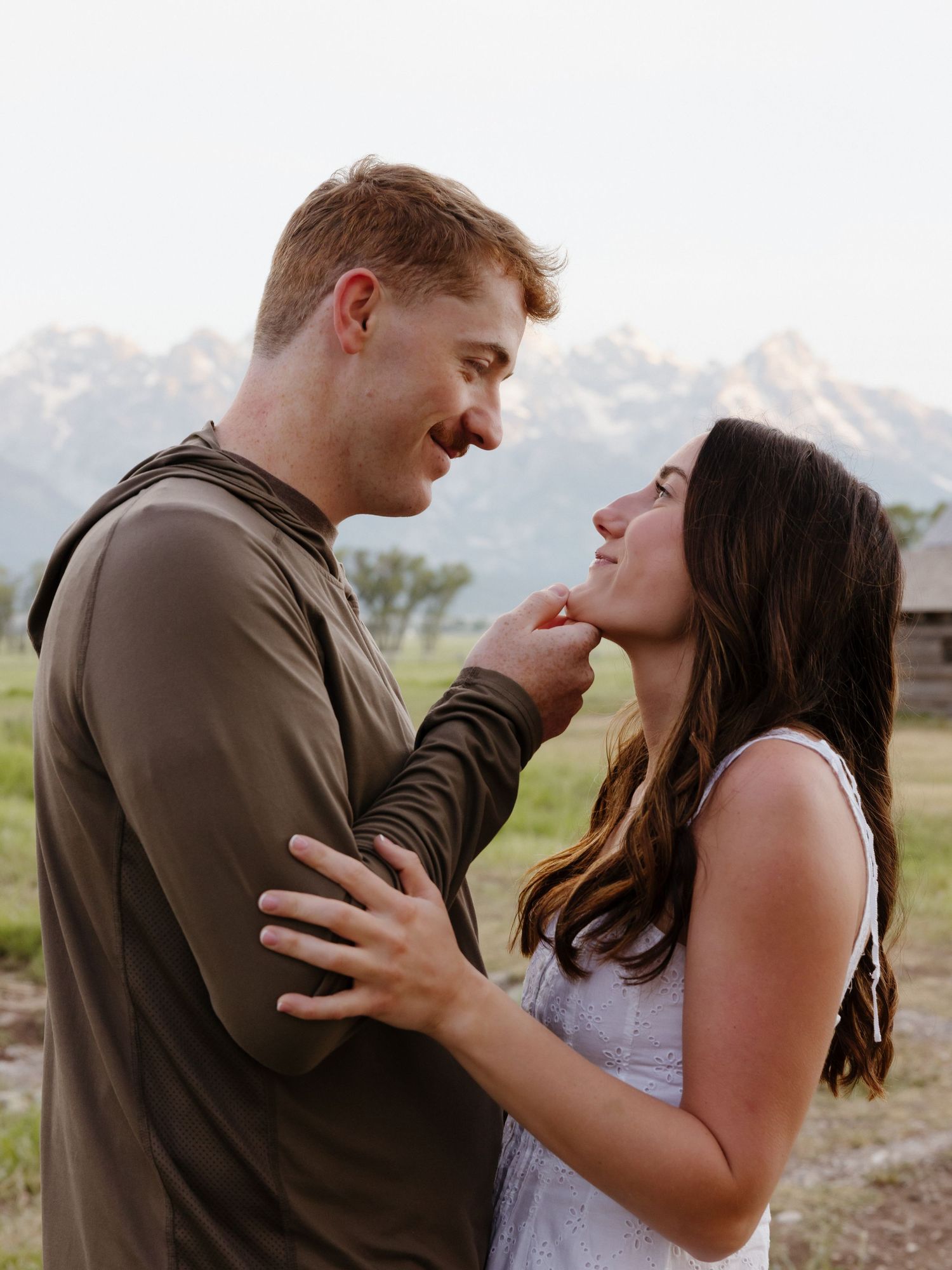 Smiling couple sharing a tender moment in a field with the Grand Teton mountains behind them.