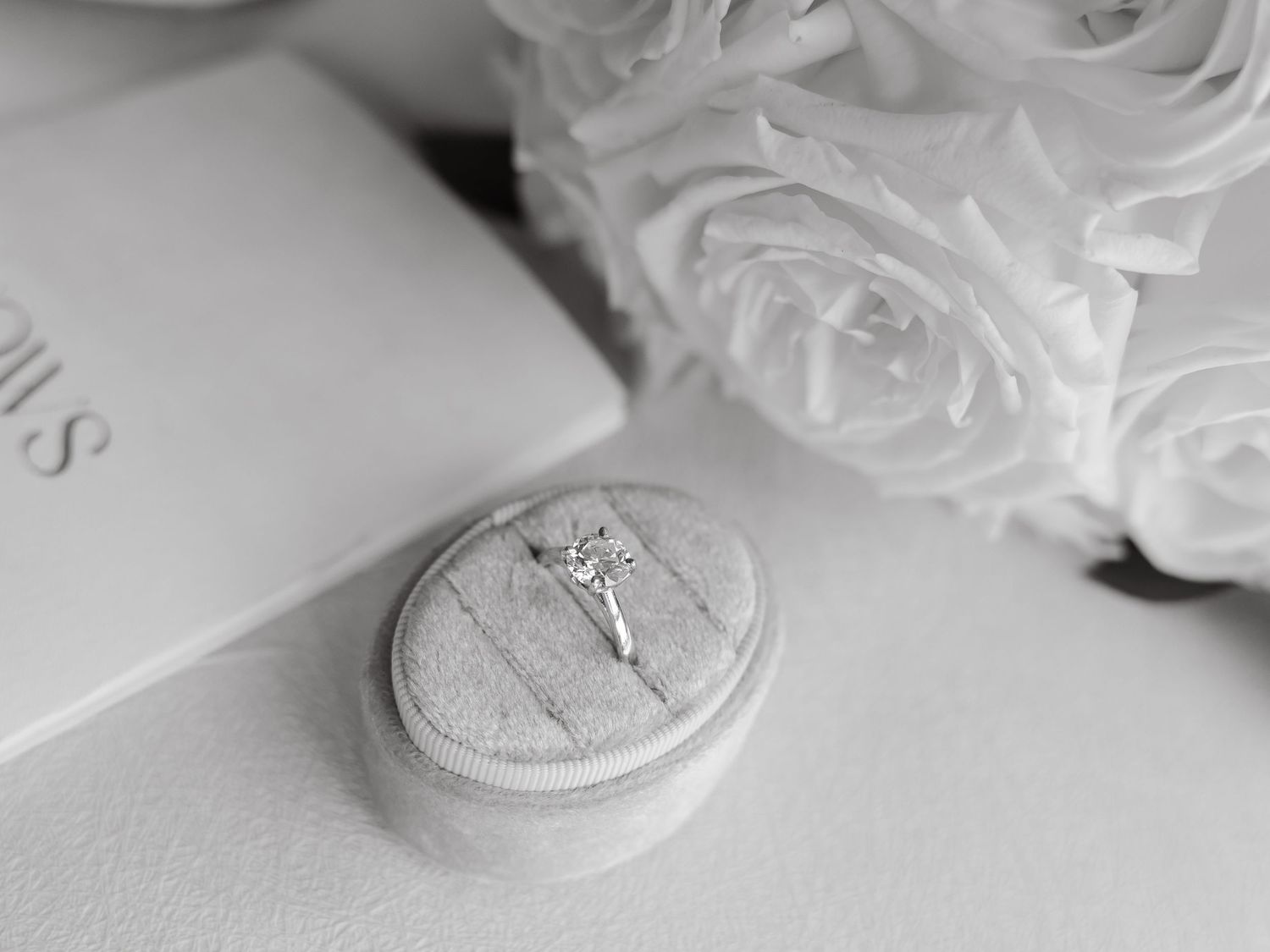Diamond solitaire engagement ring displayed in a velvet ring box beside white roses and a book.