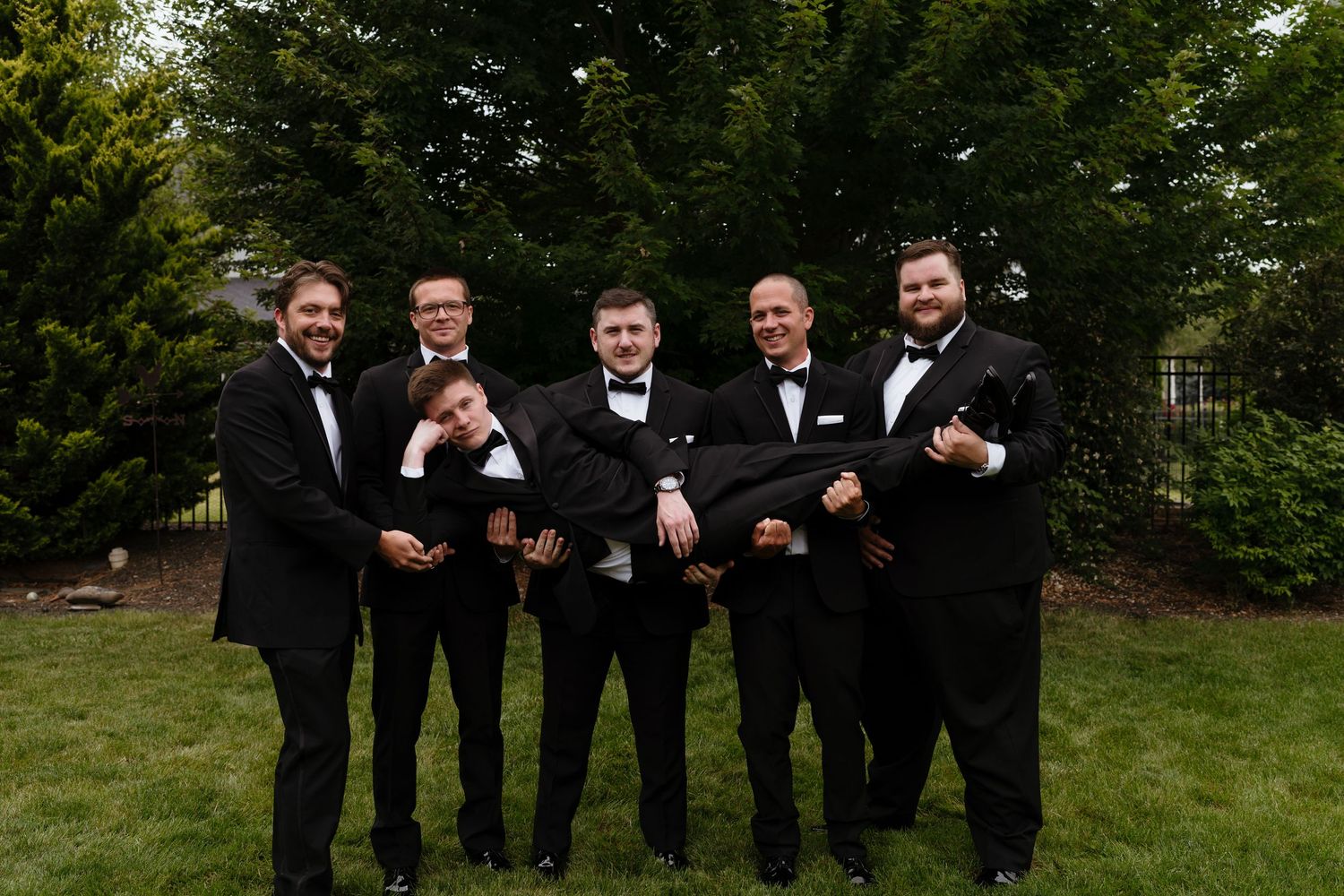 Six groomsmen in black tuxedos and bow ties pose playfully outdoors, one being held horizontally by the group.