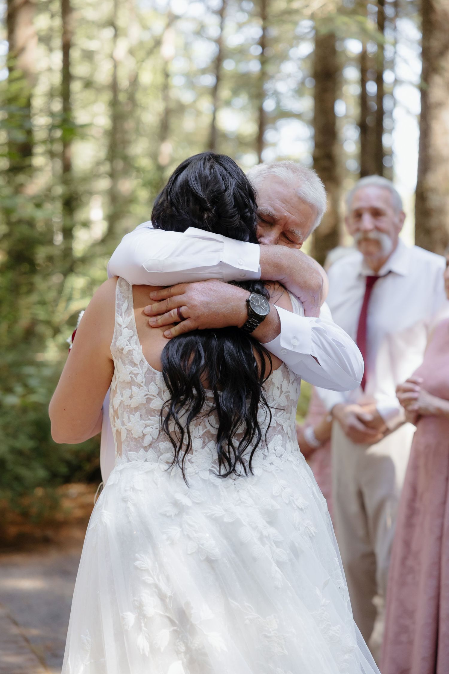 Emotional moment as father of the bride hugs his daughter in a white wedding gown at outdoor forest ceremony.