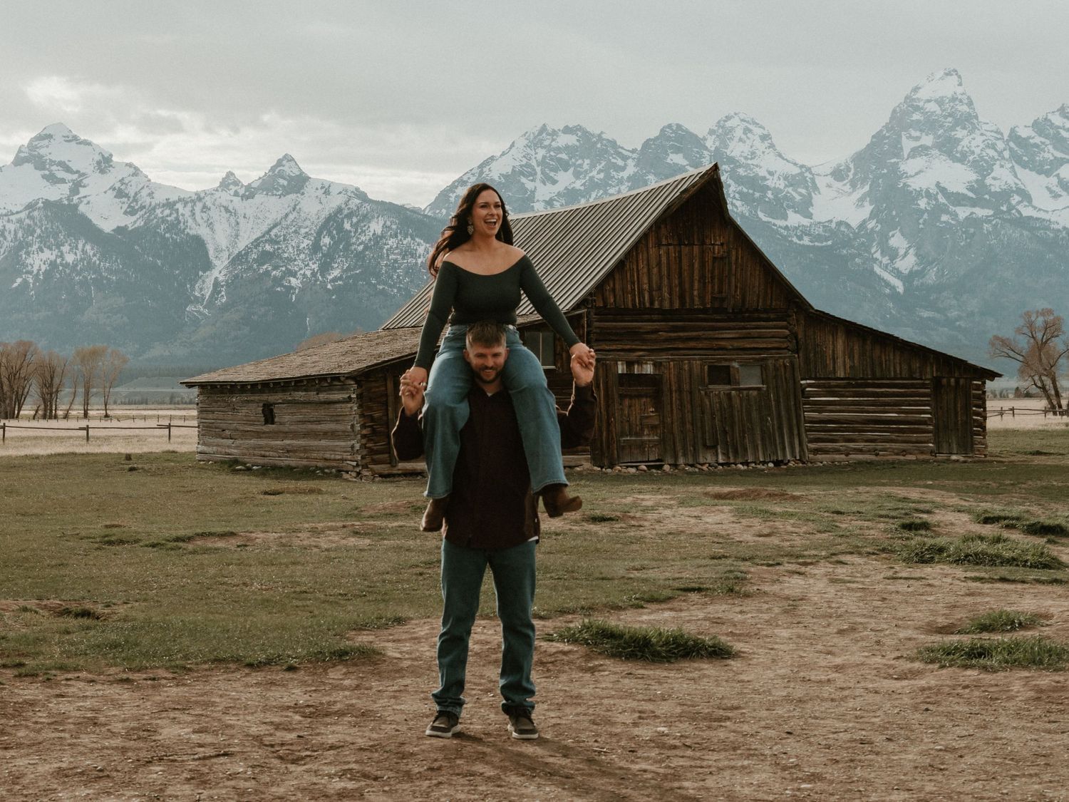 Woman sitting on mans shoulders smiling in front of a historic Mormon Row barn and Grand Teton peaks.