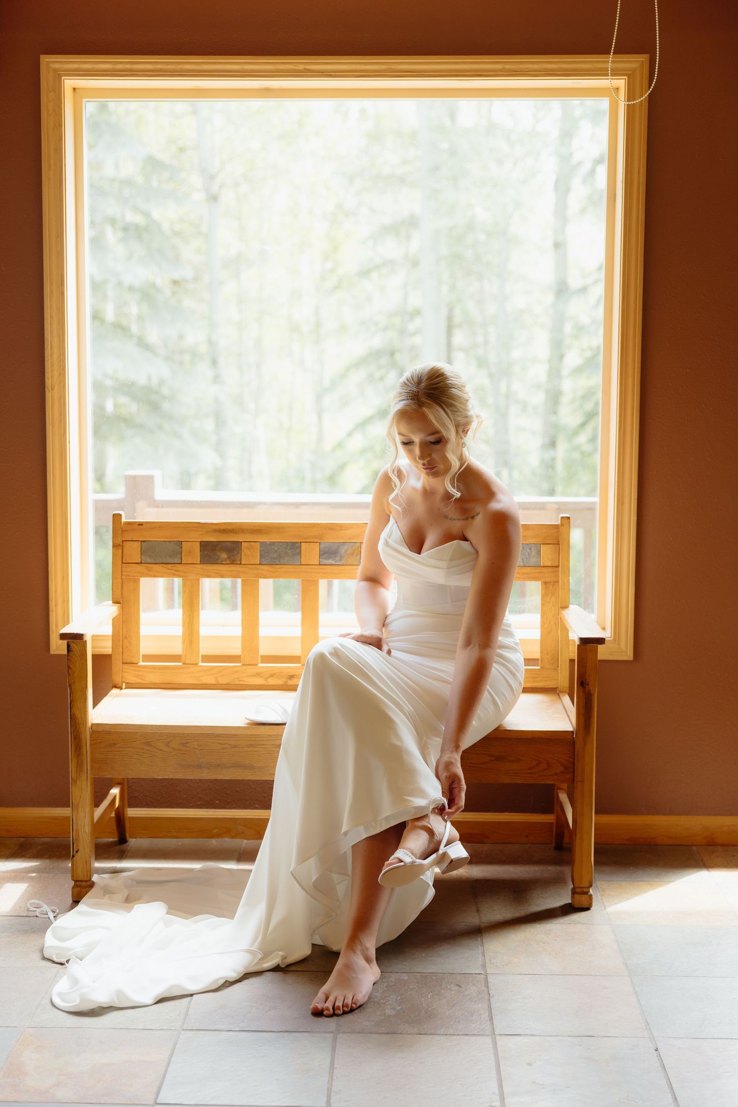 Bride in white wedding gown sitting on wooden bench by window putting on her bridal shoes.