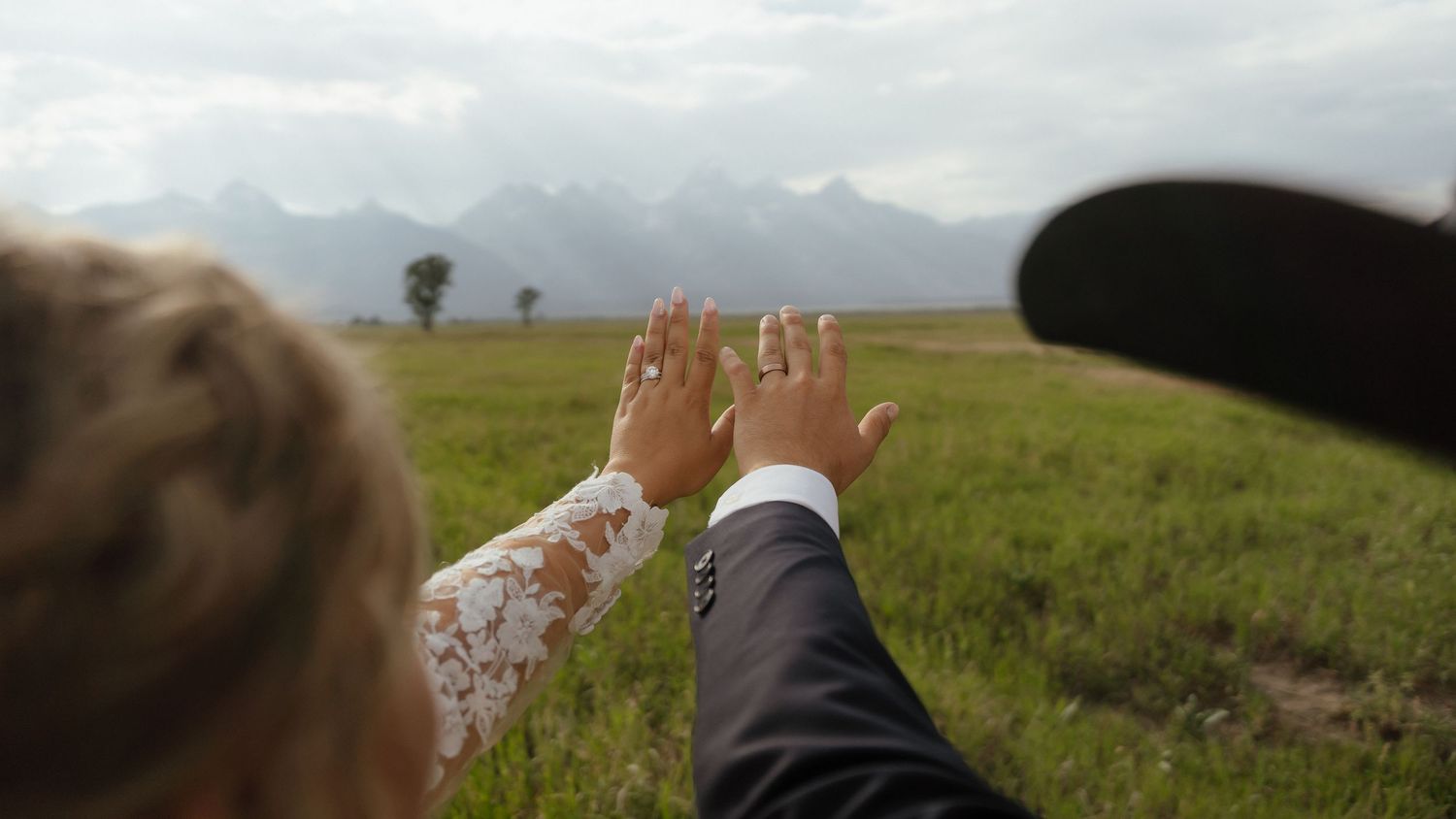 Close-up of bride and groom holding hands with wedding rings visible against a green mountain meadow.