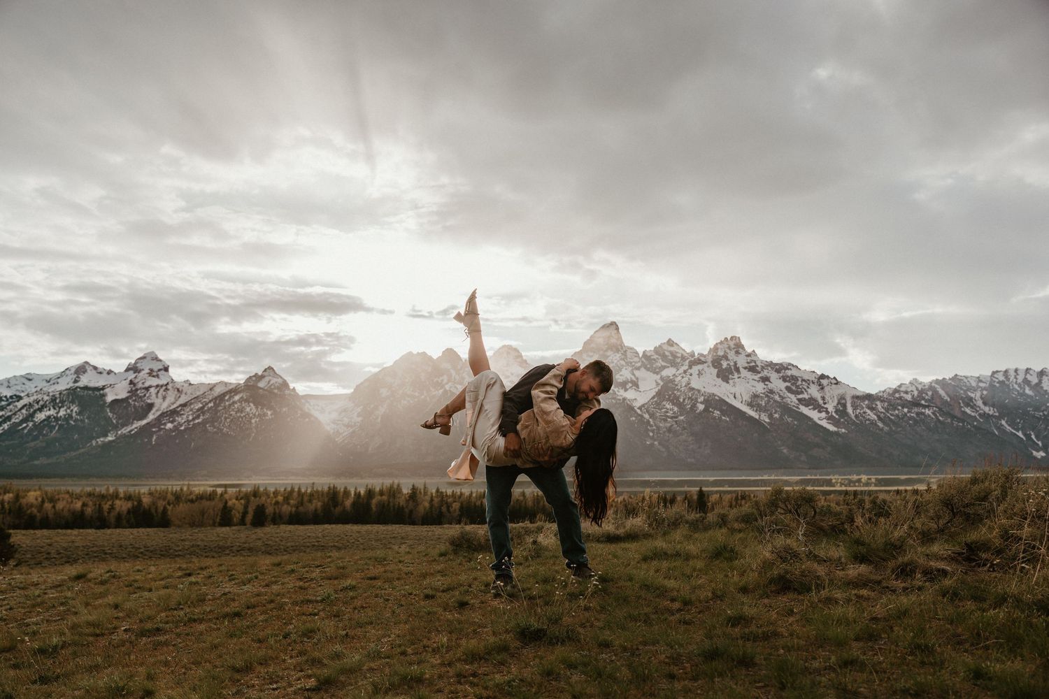 Couple dancing dramatically in a mountain meadow with snow-capped peaks and dramatic cloudy sky backdrop.