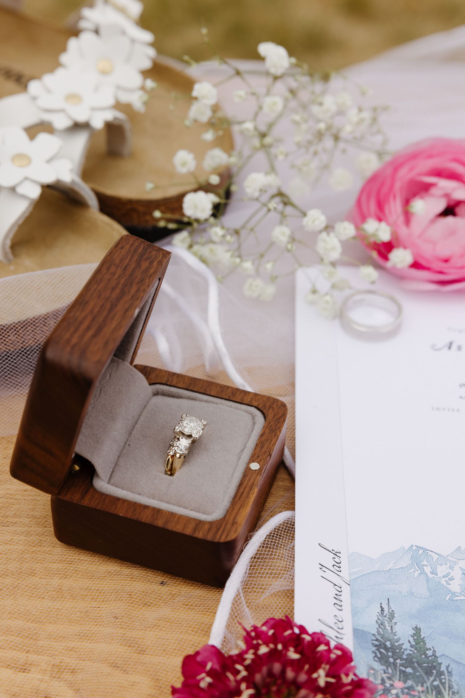Diamond engagement ring displayed in a wooden ring box surrounded by florals, veil, and wedding stationery.
