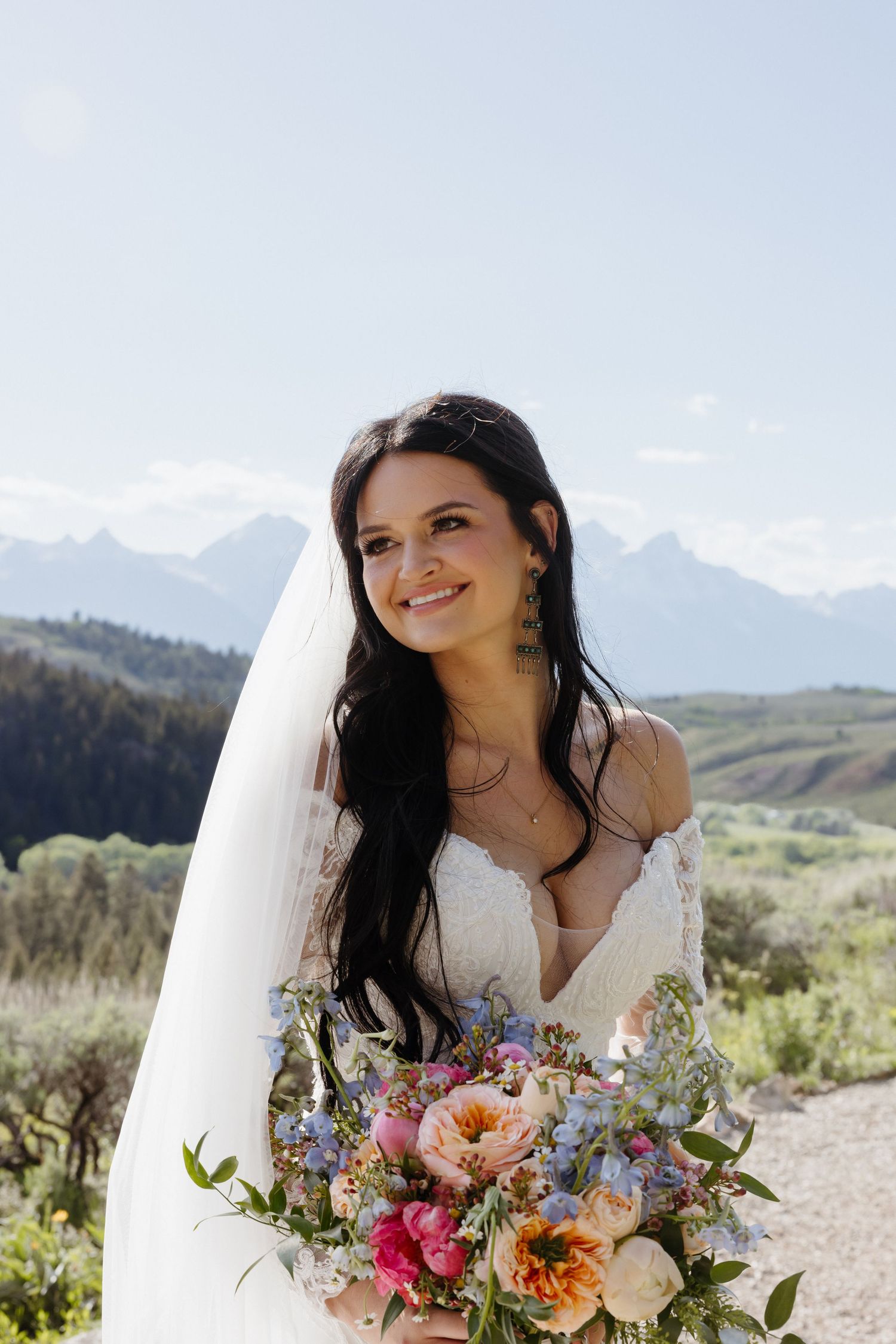 Radiant dark-haired bride in lace gown holding colorful bouquet with mountain backdrop.