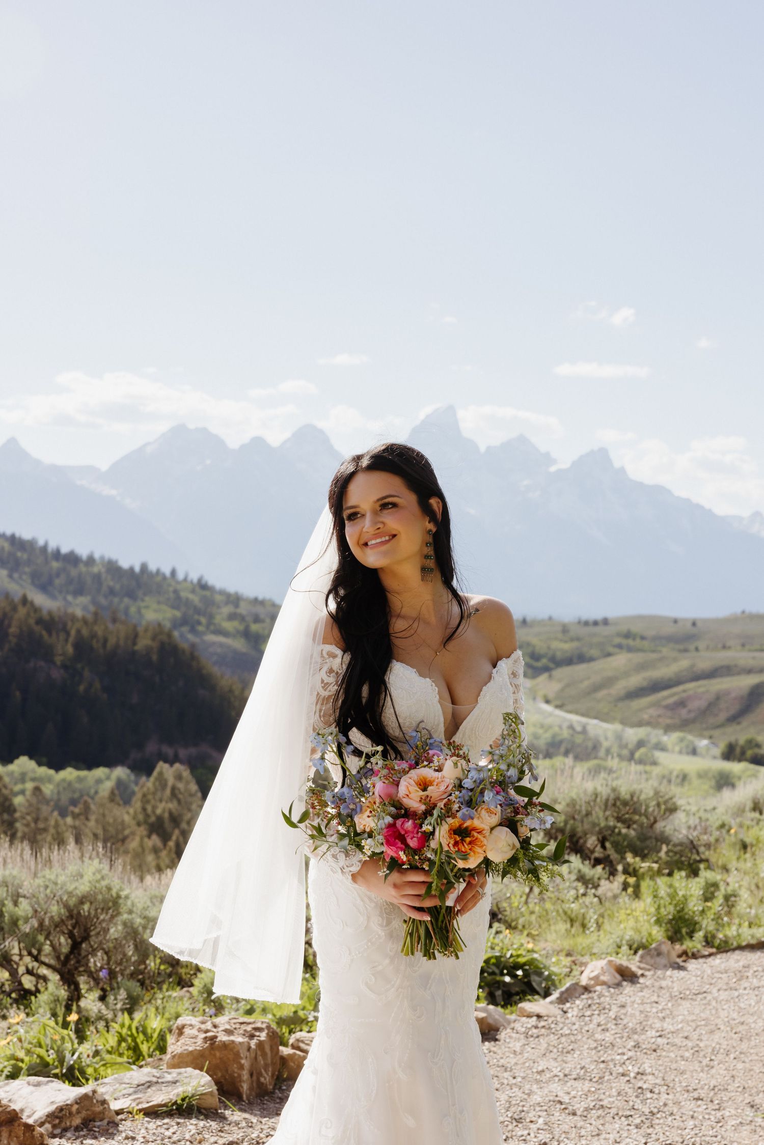 Radiant dark-haired bride holds colorful bouquet in lace gown with Grand Teton mountain backdrop.