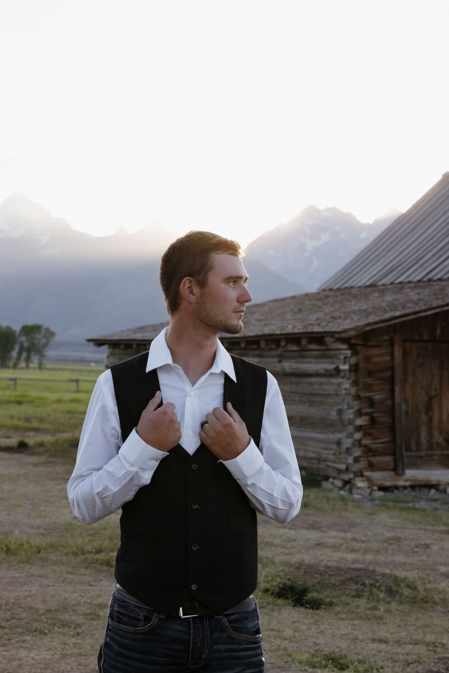 Handsome groom in white shirt and black vest adjusting attire near rustic barn with mountain scenery.
