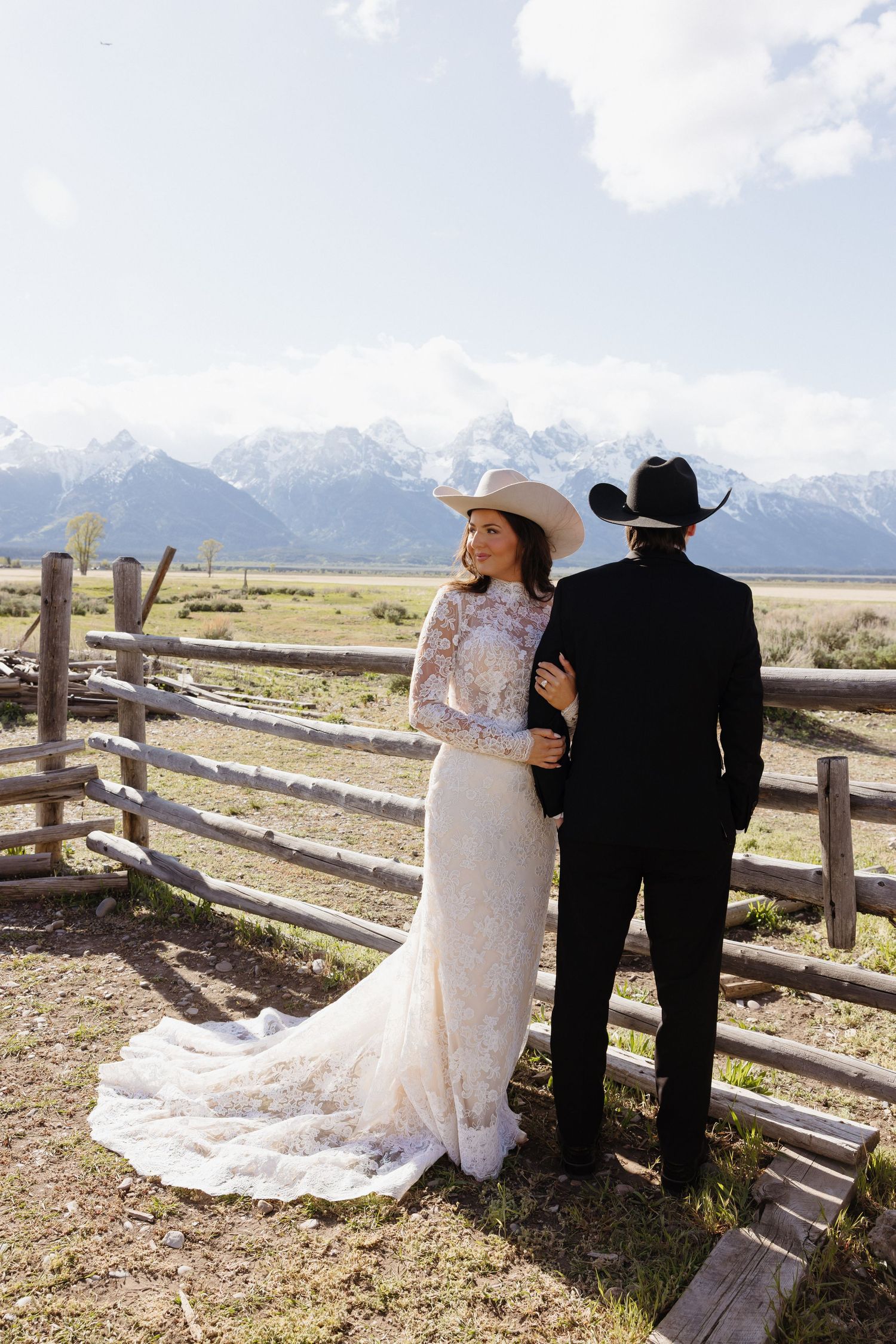 Bride and groom in cowboy hats stand by rustic wooden fence with stunning Grand Teton mountain range behind them.