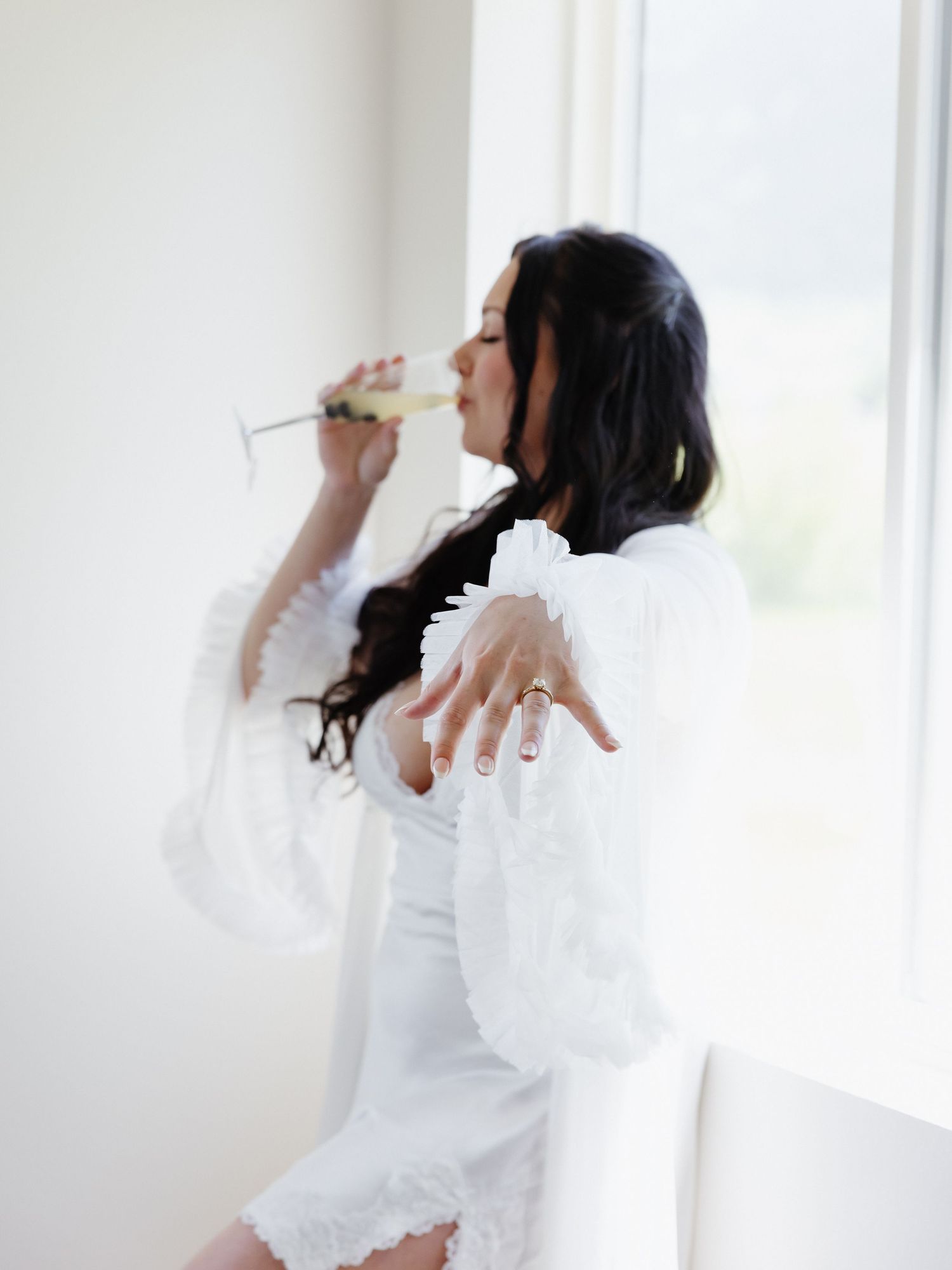 Bride in white lace robe sipping champagne by a bright window, showcasing her engagement ring.