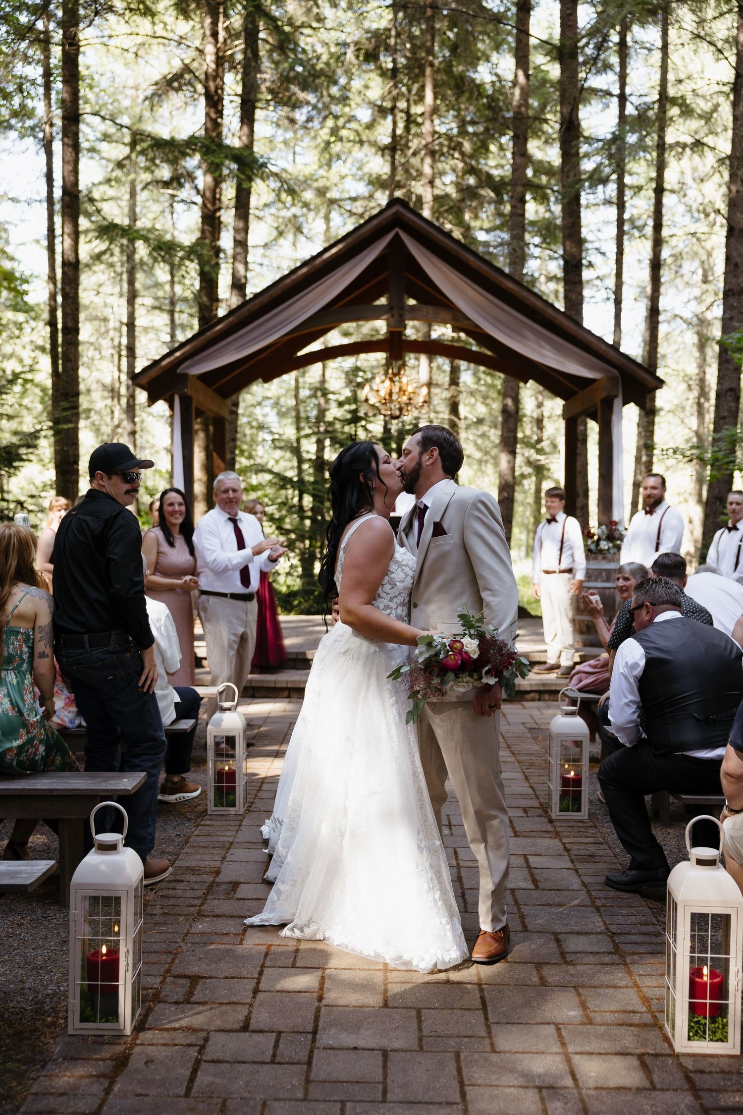 Bride and groom share first kiss at outdoor forest wedding ceremony beneath wooden pergola with guests watching on both sides.