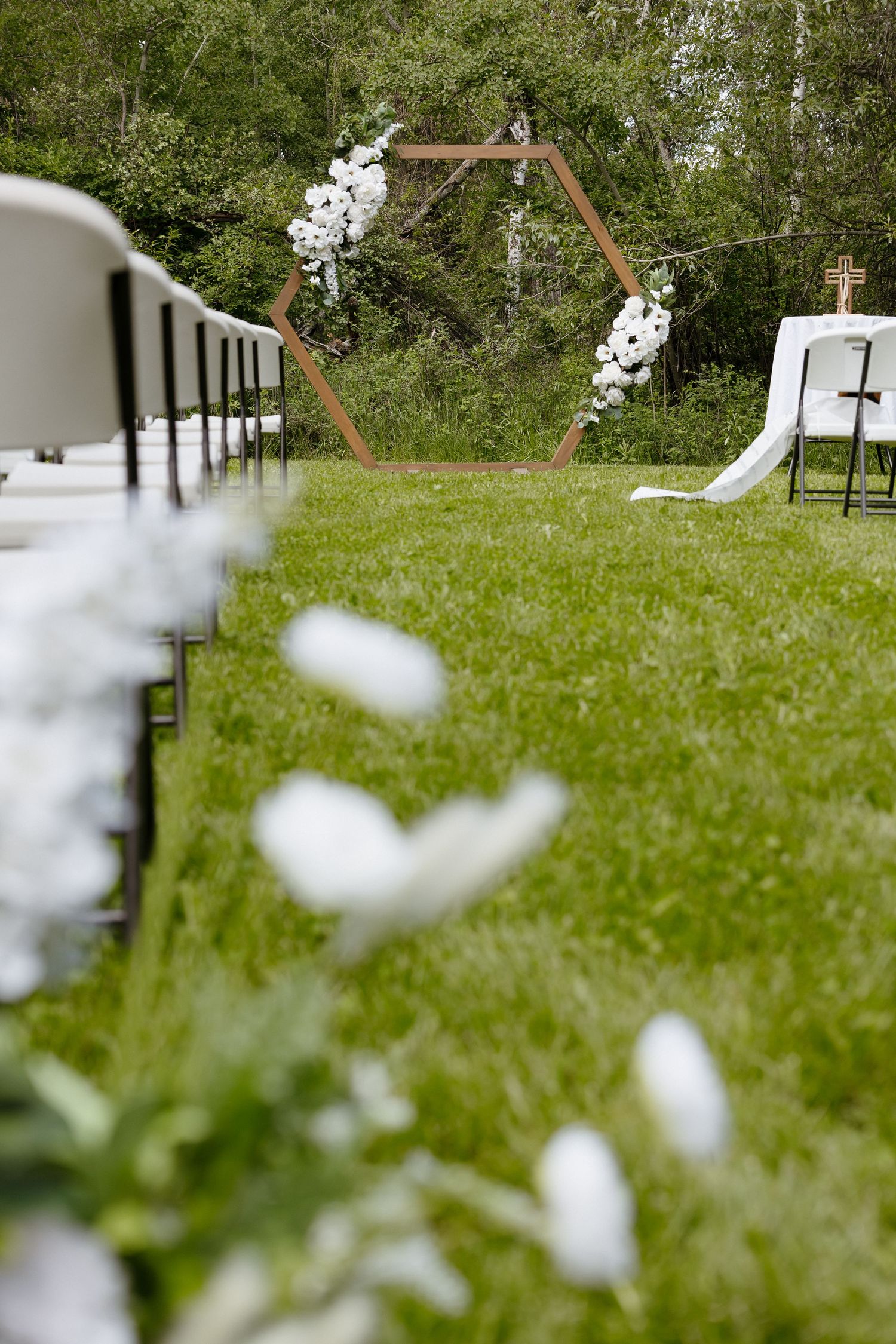 Hexagonal wooden wedding arch with white flowers on green grass outdoor venue.