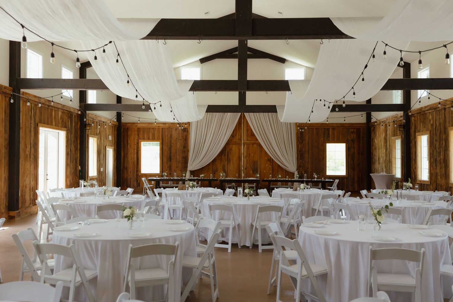 Rustic barn wedding reception venue with white draped ceiling, string lights, and round white-clothed tables.