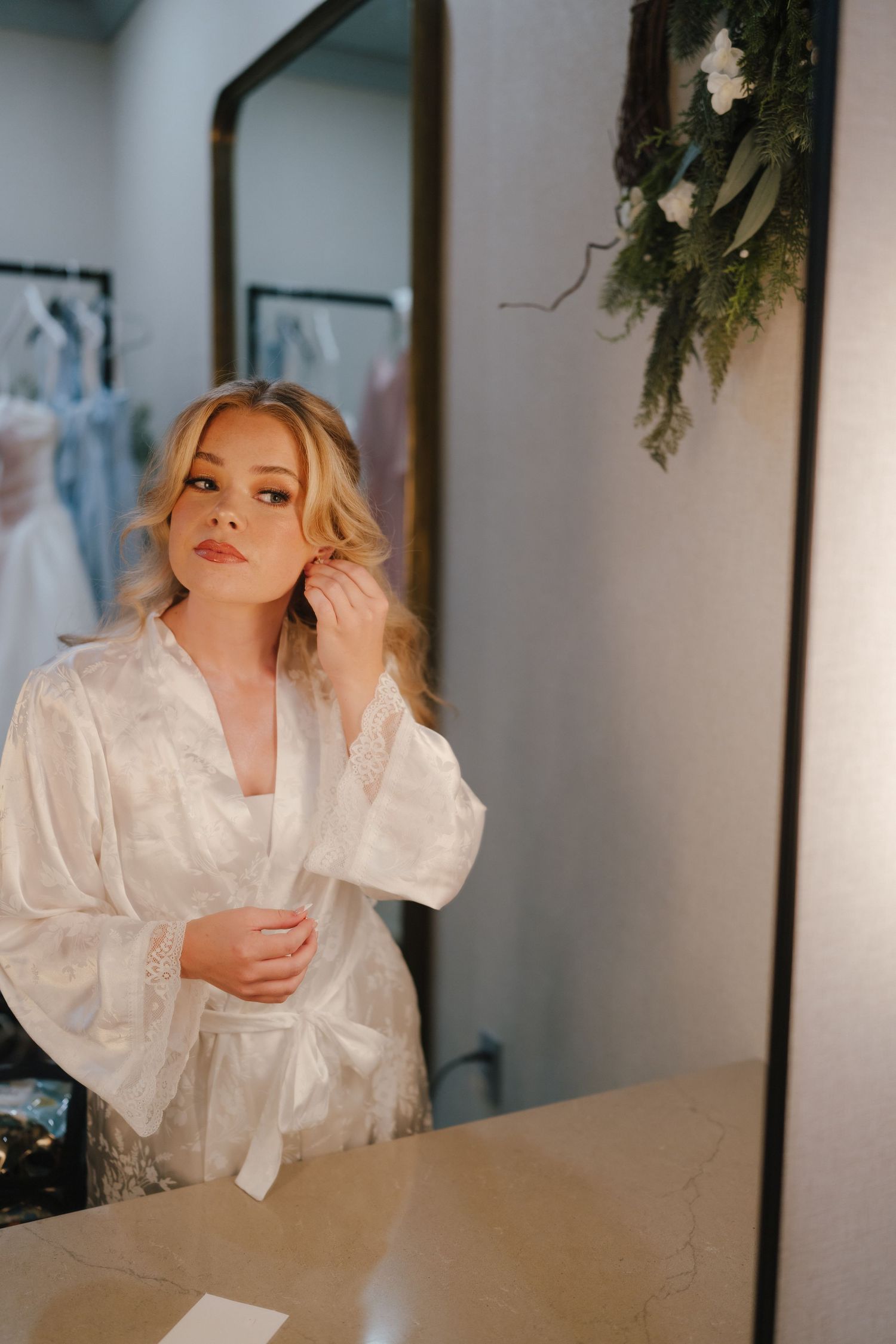 Blonde bride in white satin robe touching her ear, standing before a mirror in a bridal boutique.
