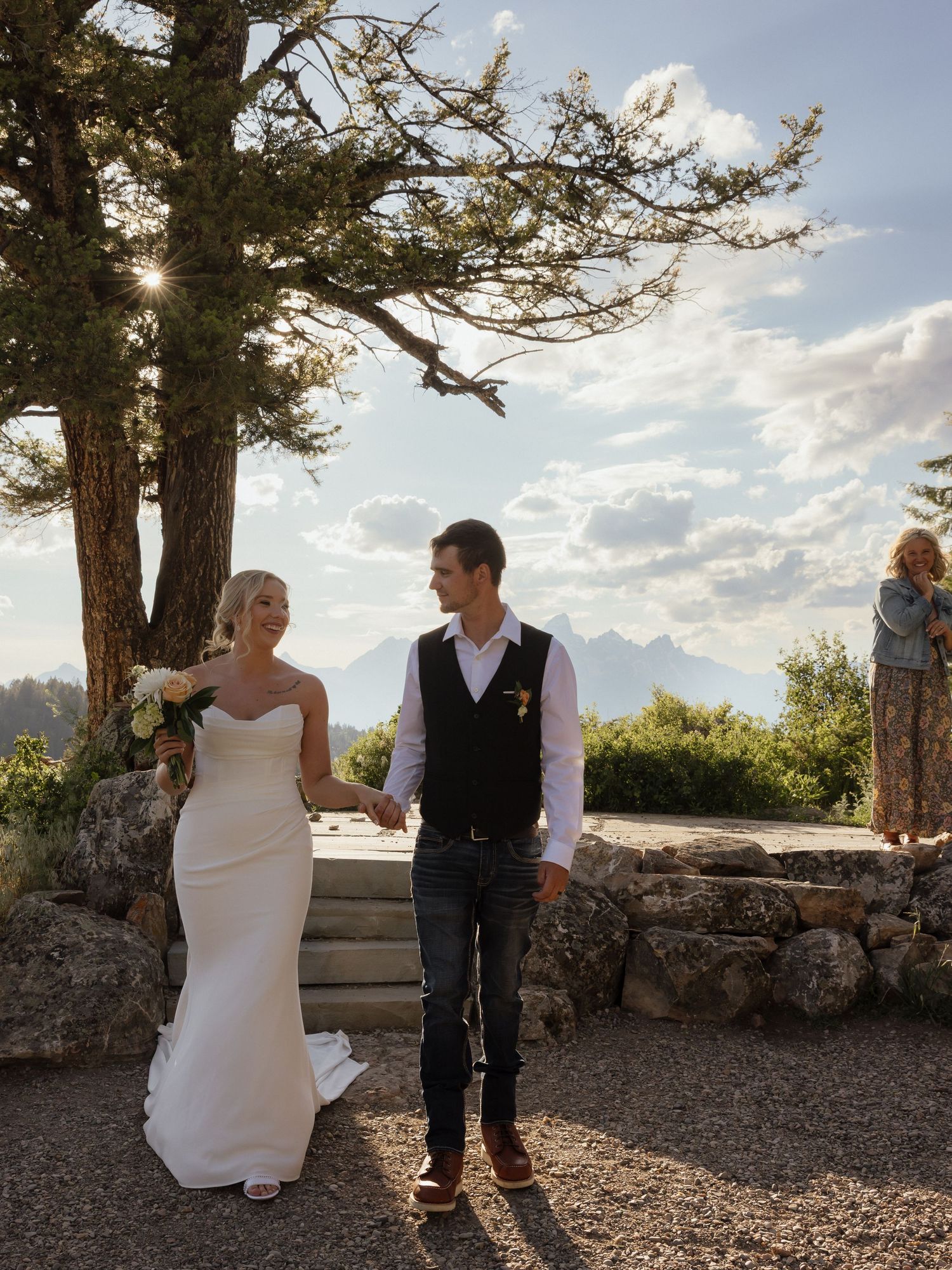Bride in white gown and groom in vest hold hands at scenic outdoor wedding venue with Teton views.