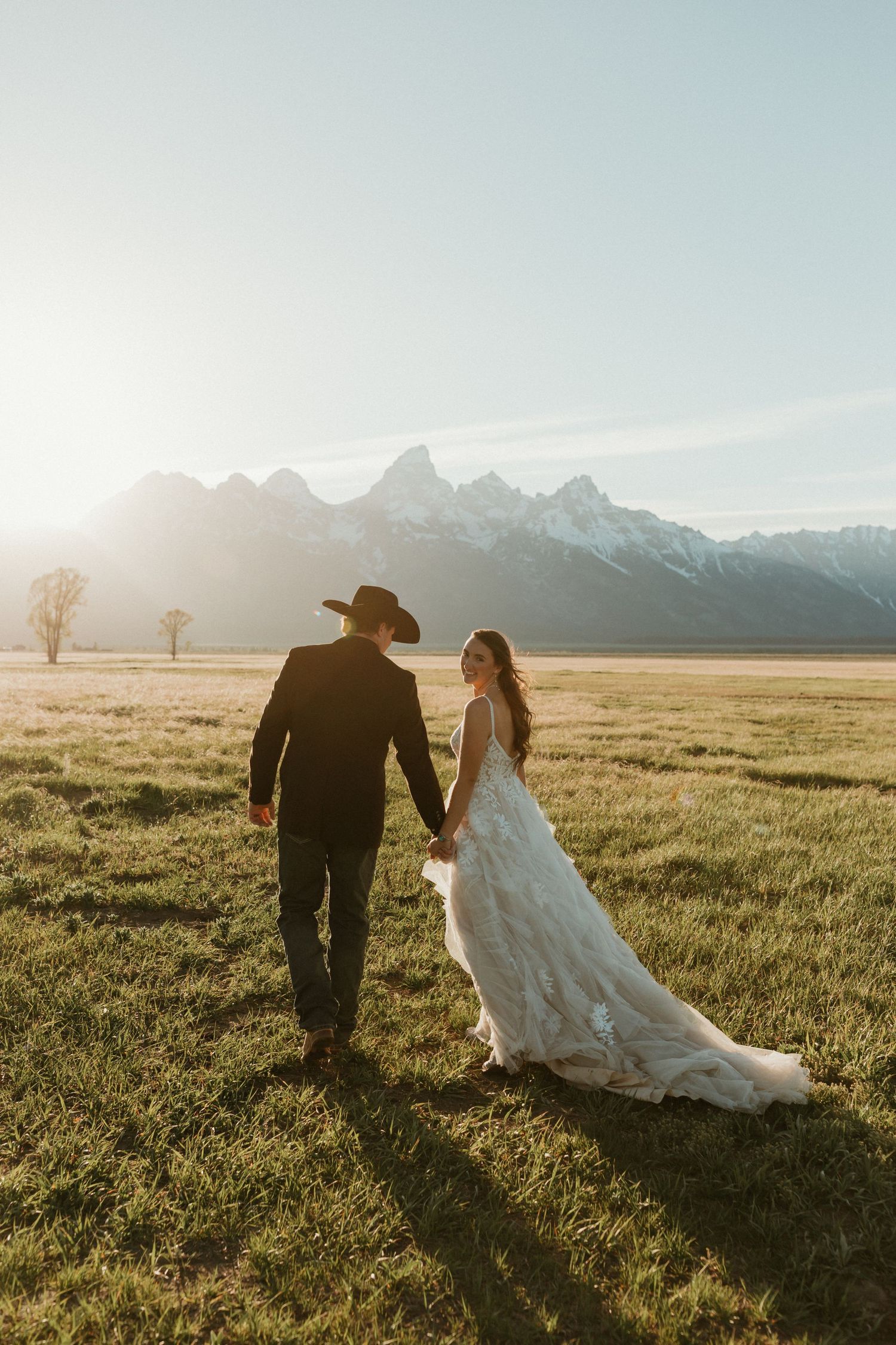 Bride and groom walking hand in hand through a sunlit meadow with the Grand Teton mountains in the background.
