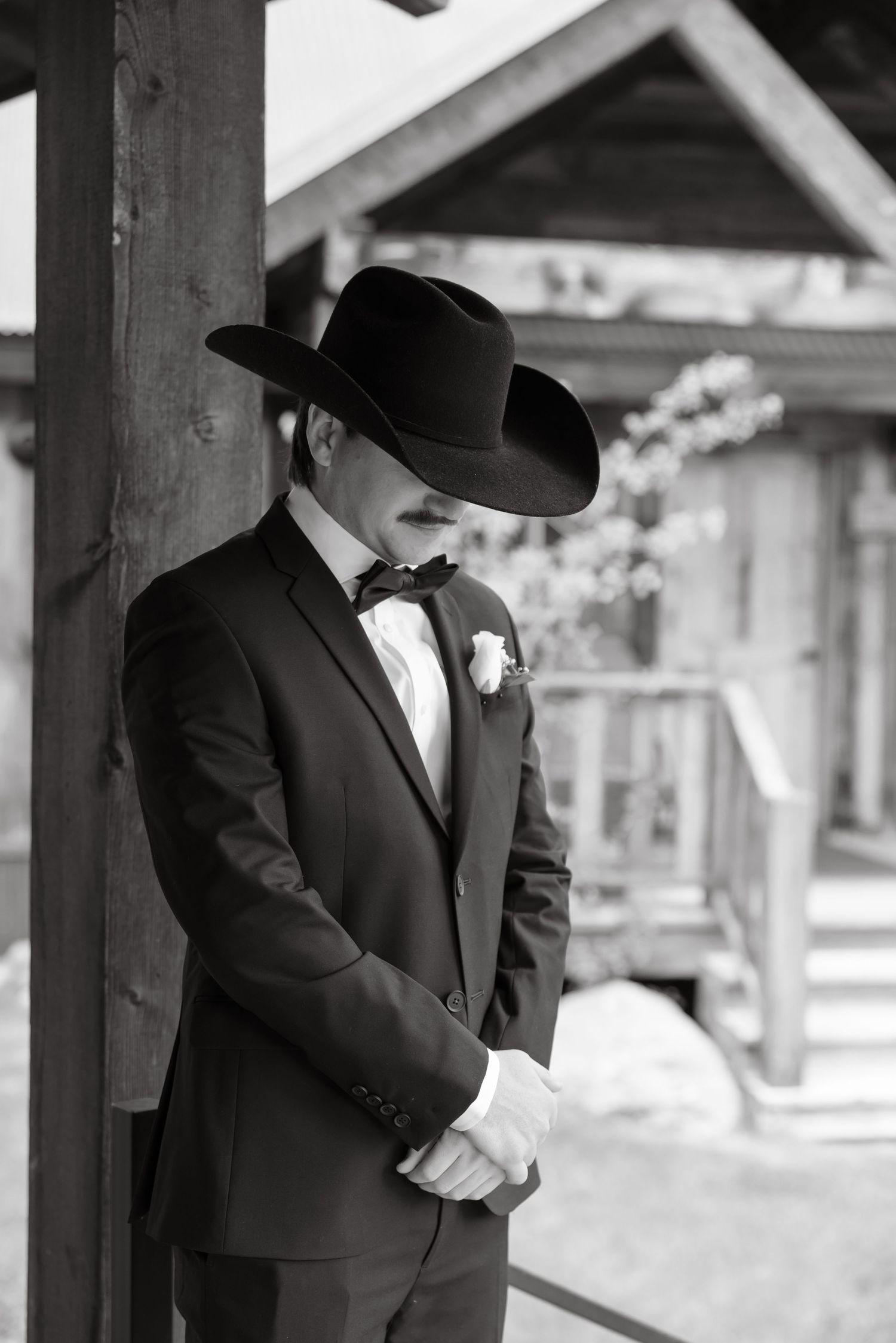 Black and white photo of groom in black tuxedo and cowboy hat with boutonniere at rustic venue.