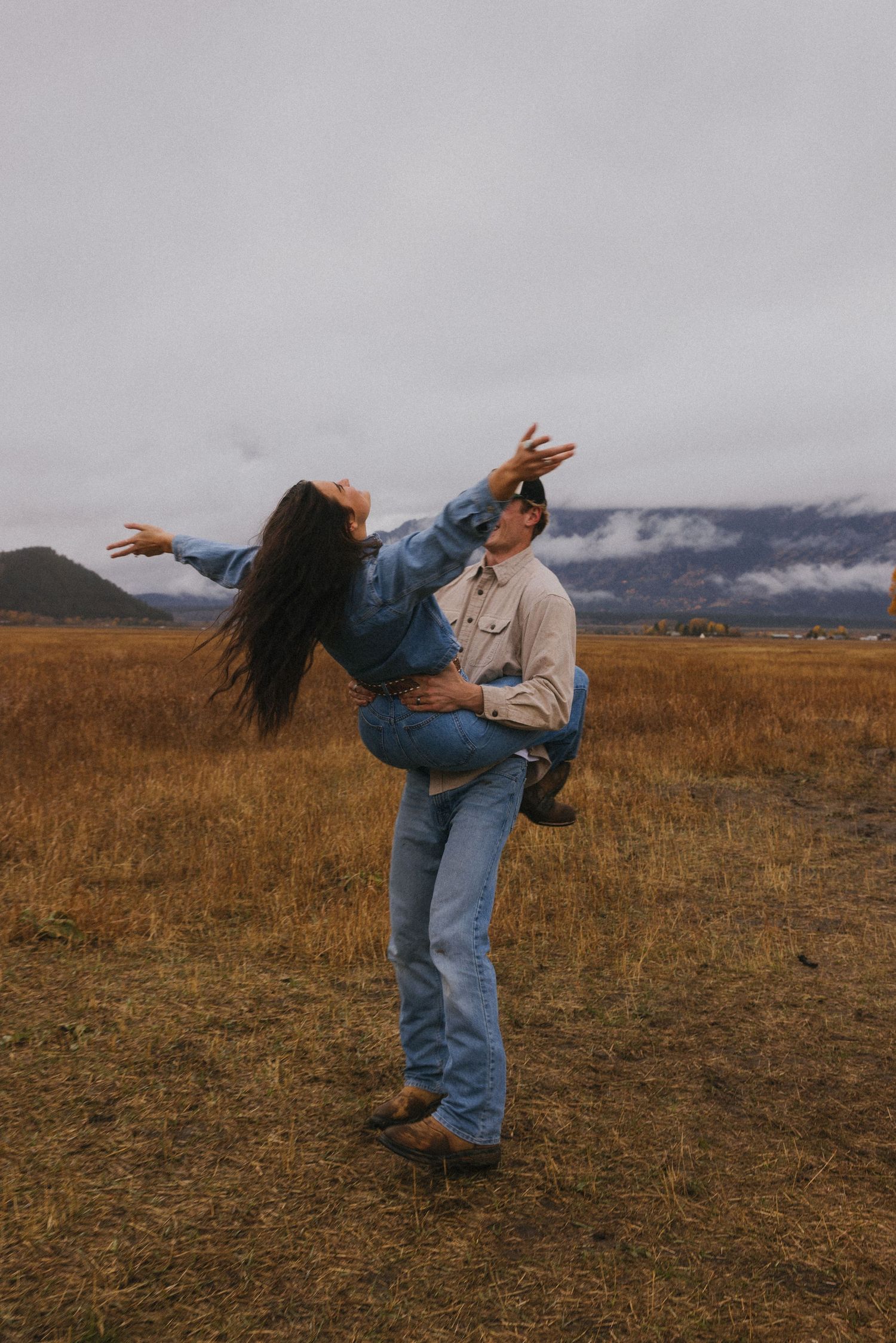 Man lifting woman in a joyful pose in a golden field with misty mountain backdrop.
