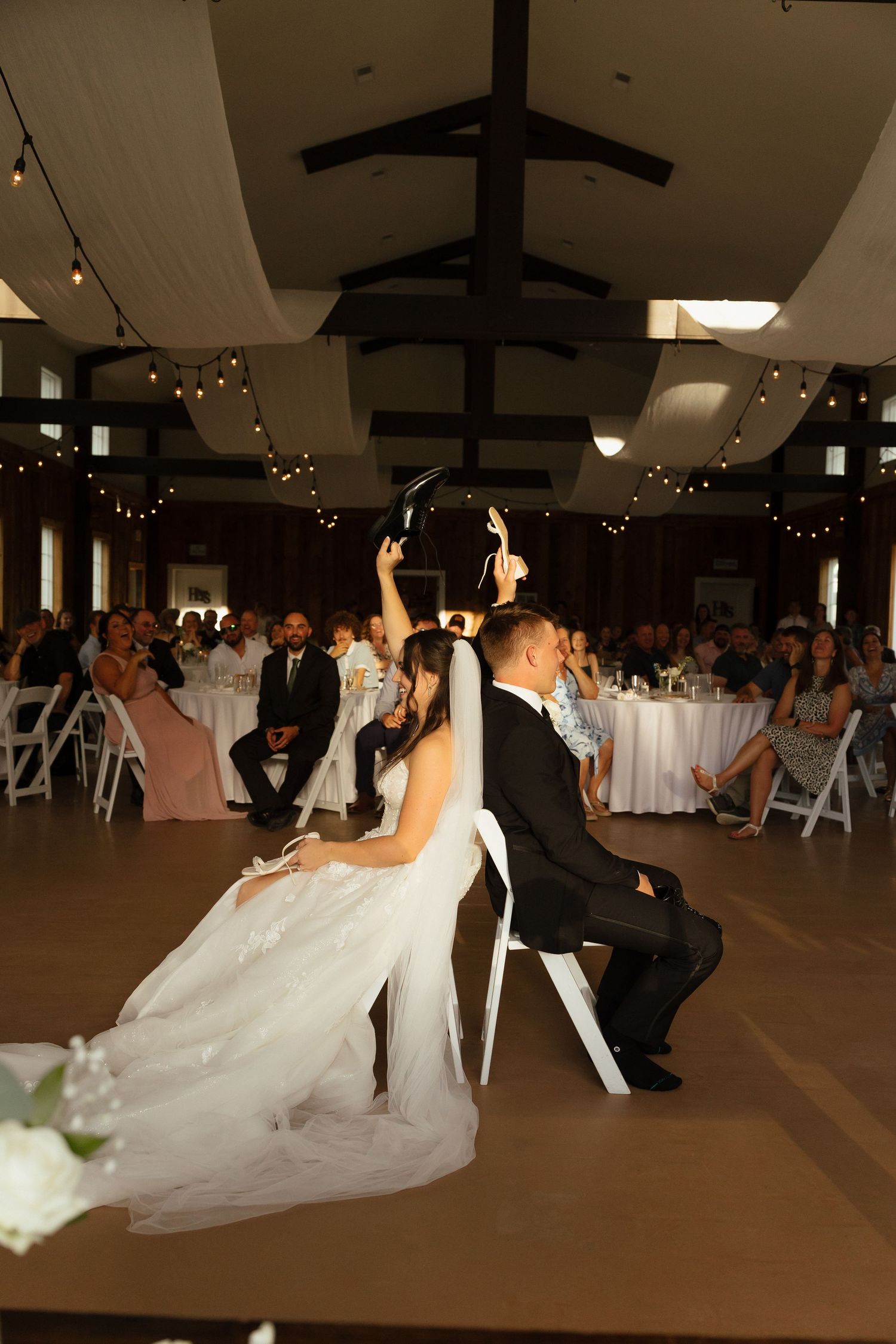 Bride and groom play wedding game seated in chairs on dance floor before guests.
