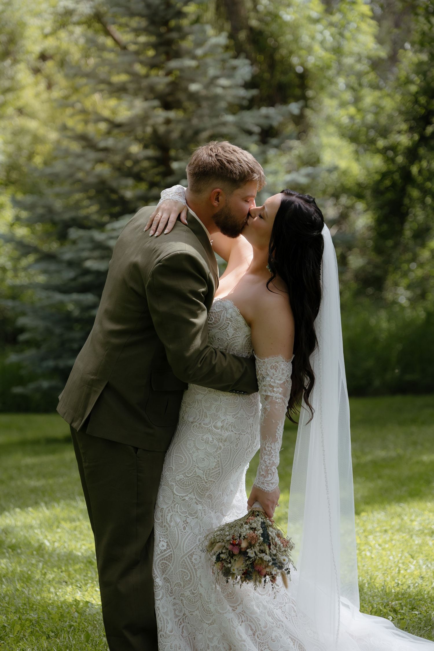 Bride and groom share a romantic kiss outdoors, bride holding a bouquet in a lace wedding gown.