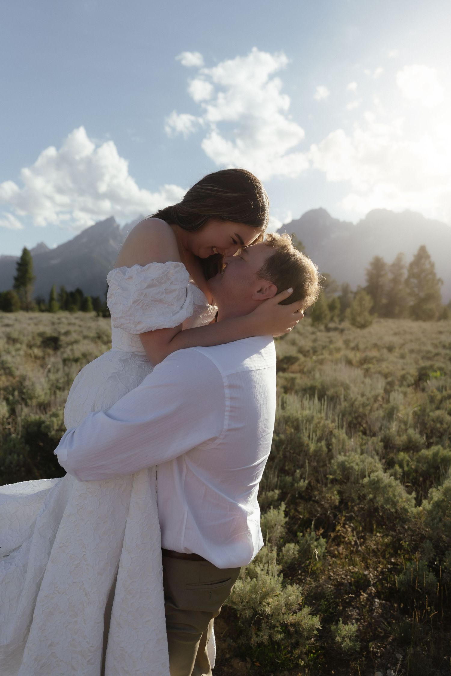 Groom lifts bride in white off-shoulder dress as they share romantic moment in sunlit Grand Teton meadow.