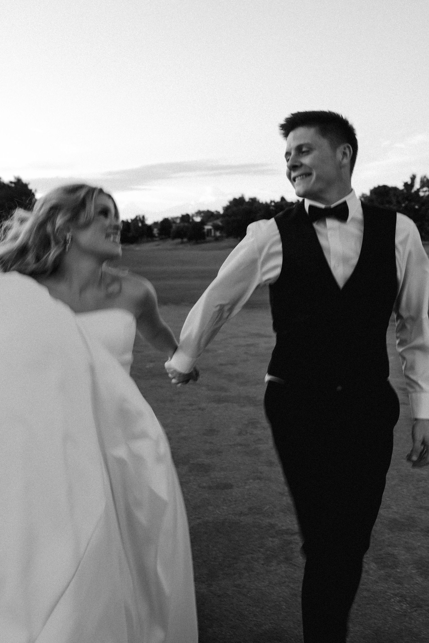 Black and white photo of a smiling bride and groom holding hands while walking outdoors on their wedding day.