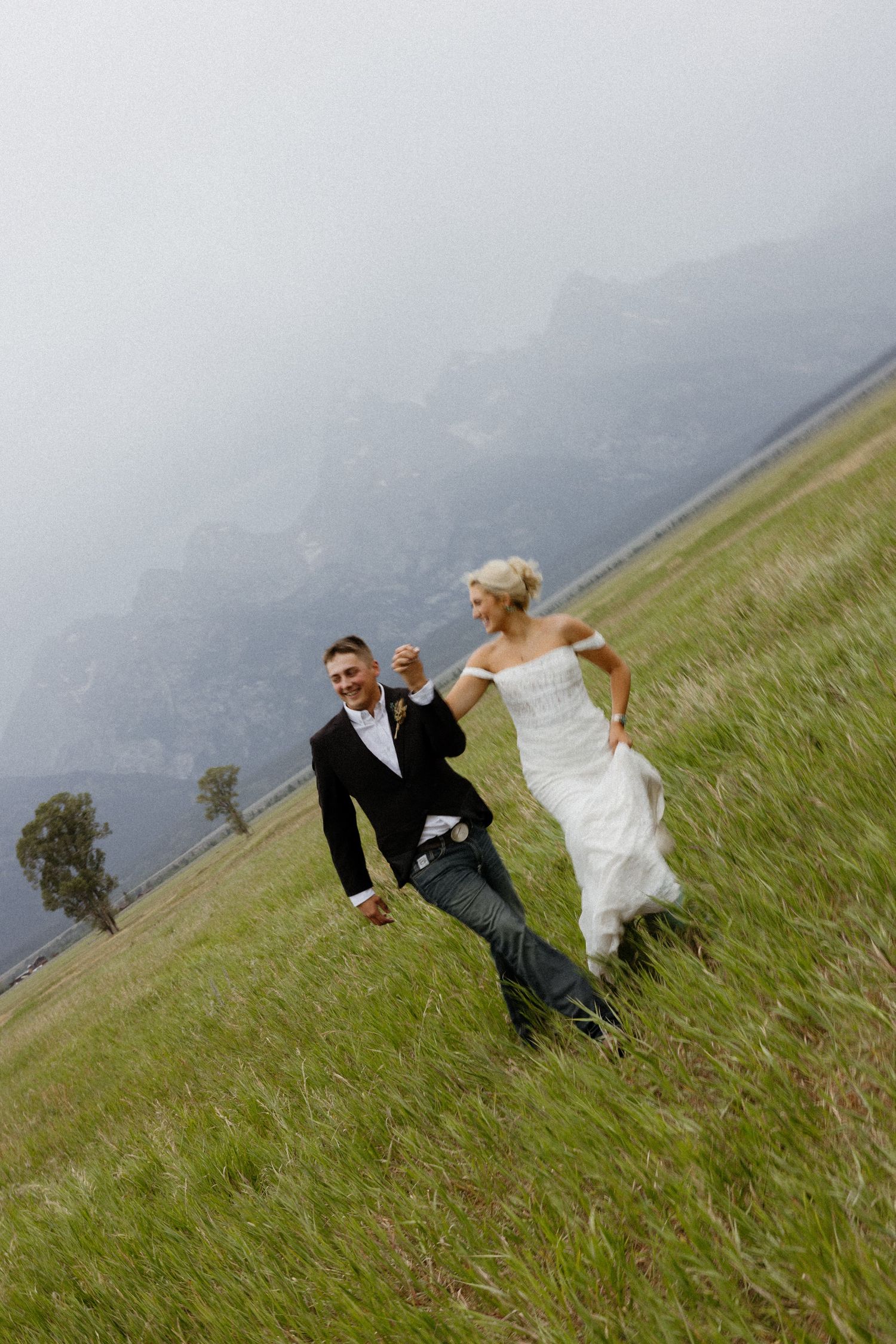 Bride and groom running joyfully through green meadow with misty mountains backdrop on their wedding day.