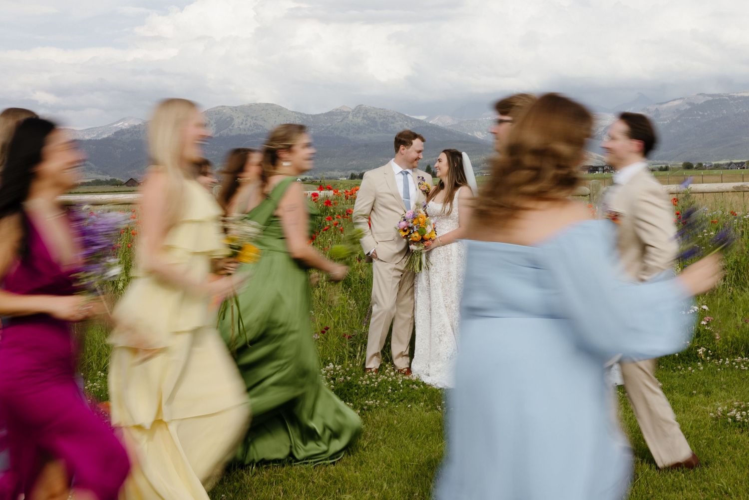 Blurred motion shot of colorful bridesmaids rushing toward bride and groom in a wildflower mountain meadow.