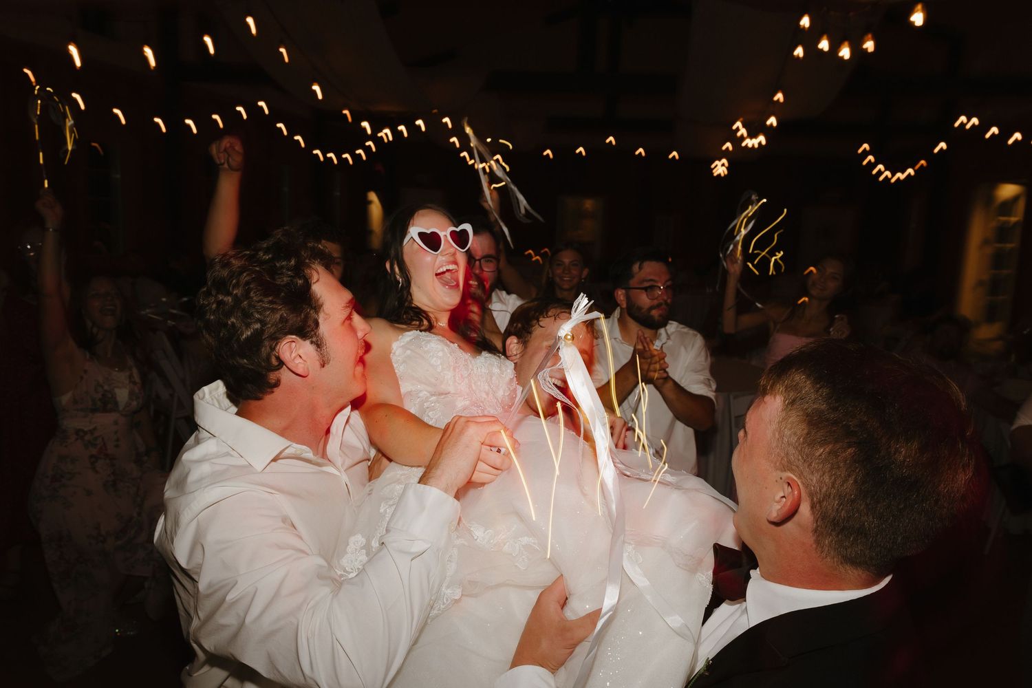 Bride wearing heart sunglasses lifted by groomsmen during sparkler wedding send-off celebration.
