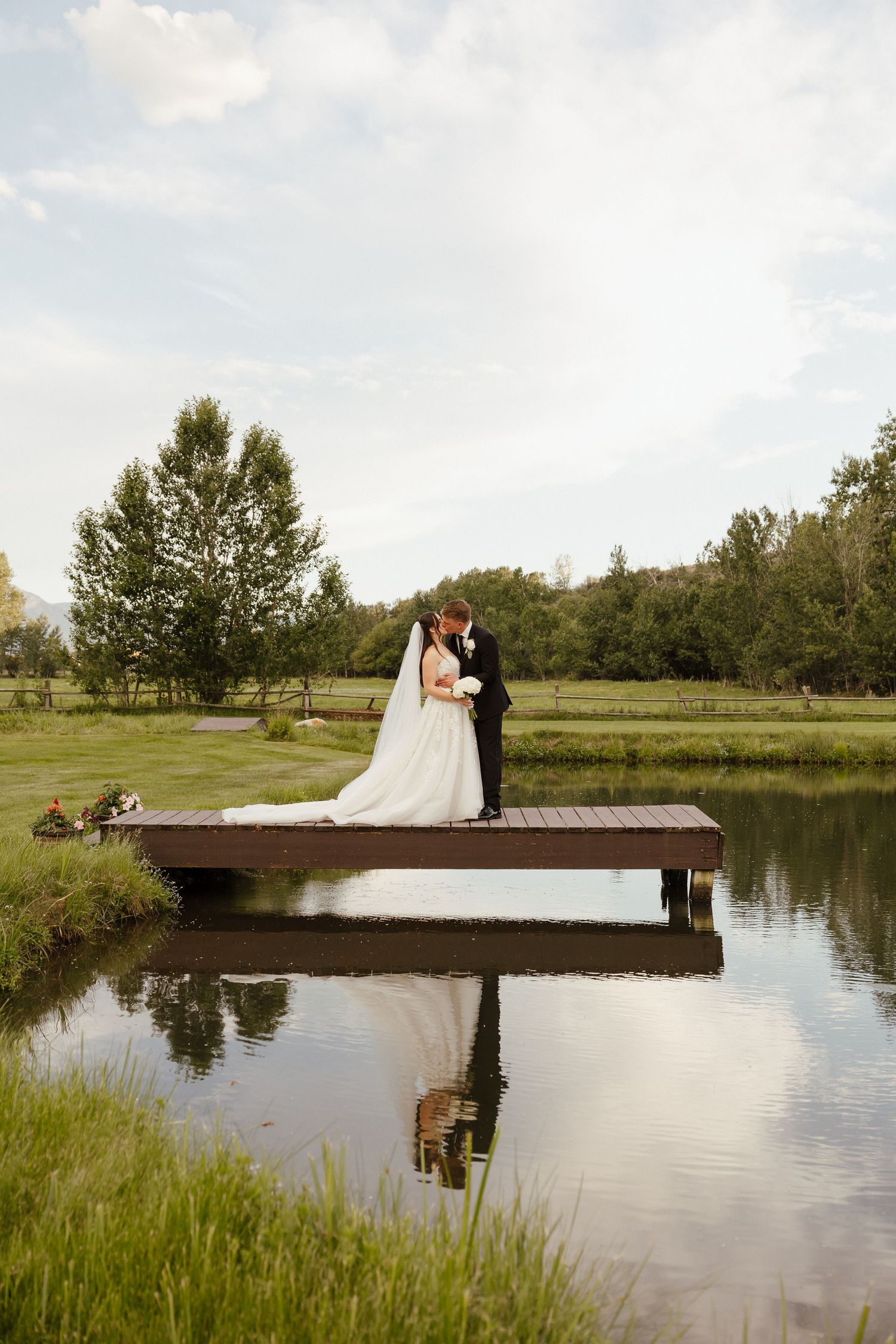 Bride and groom share a romantic kiss on a wooden dock reflected in a calm pond surrounded by lush greenery.