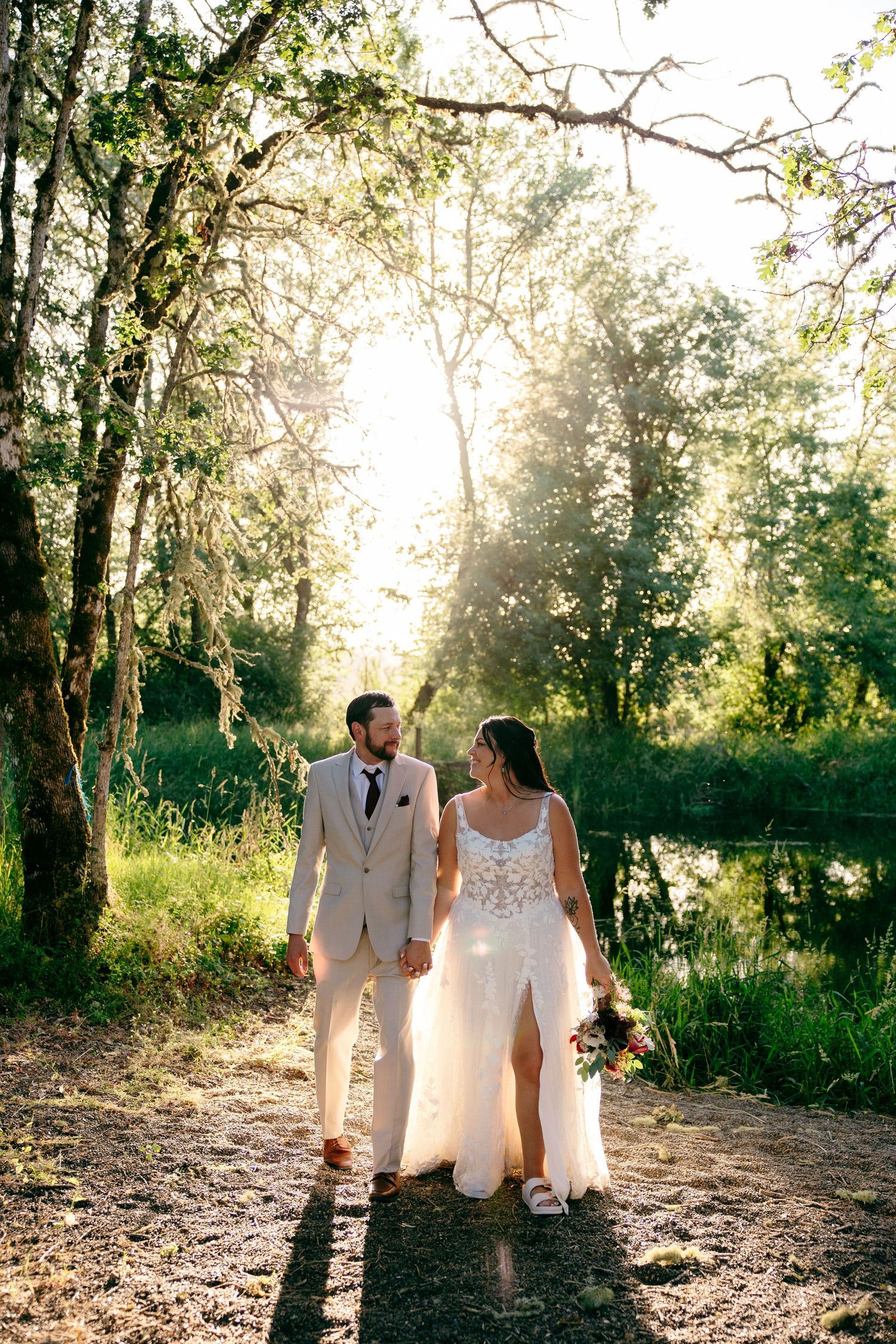 Bride and groom walking hand in hand along sunlit forest path beside a peaceful pond during wedding portraits.