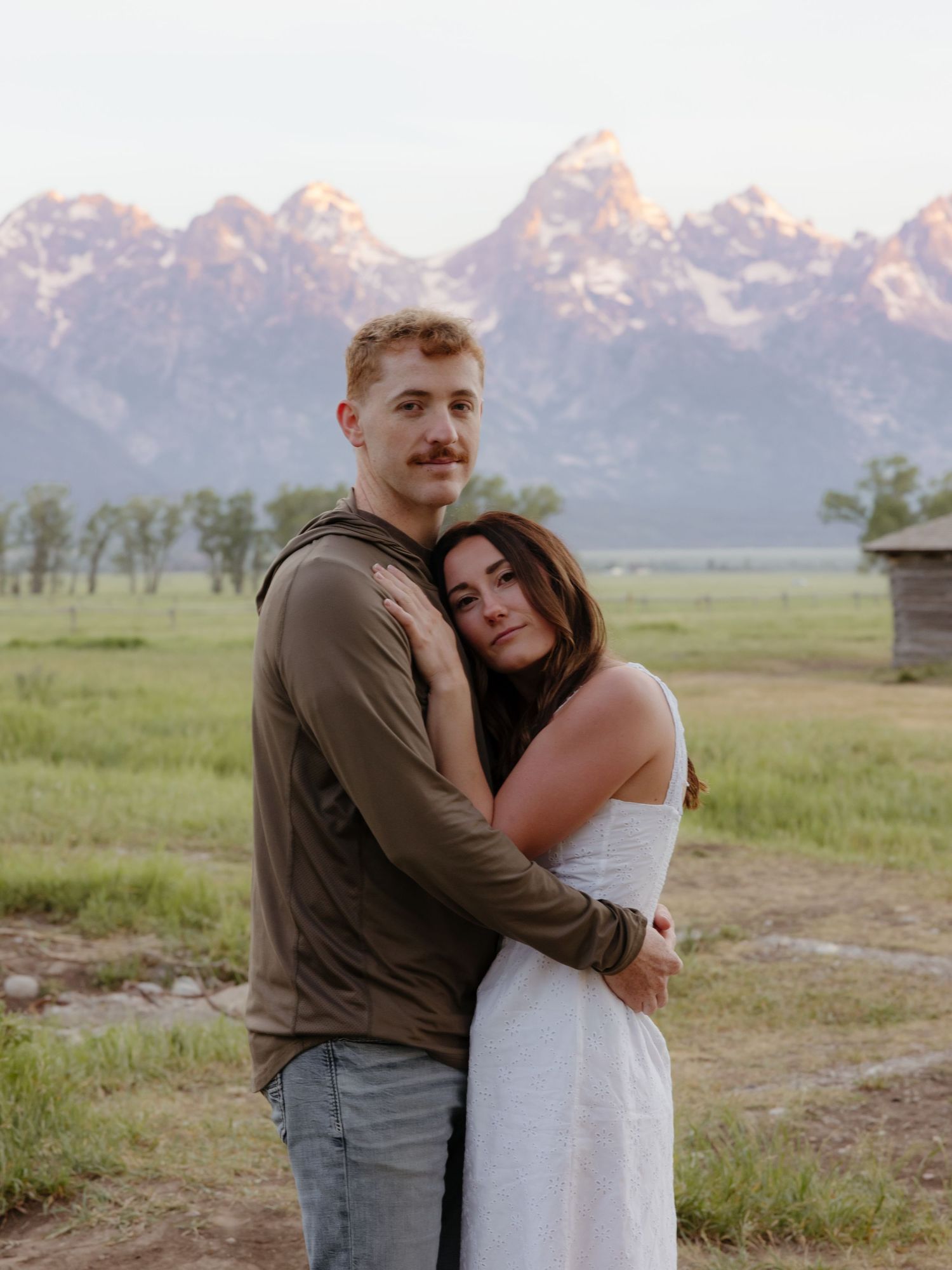 Couple embracing in a green meadow with the Grand Teton mountain range in the background at sunset.
