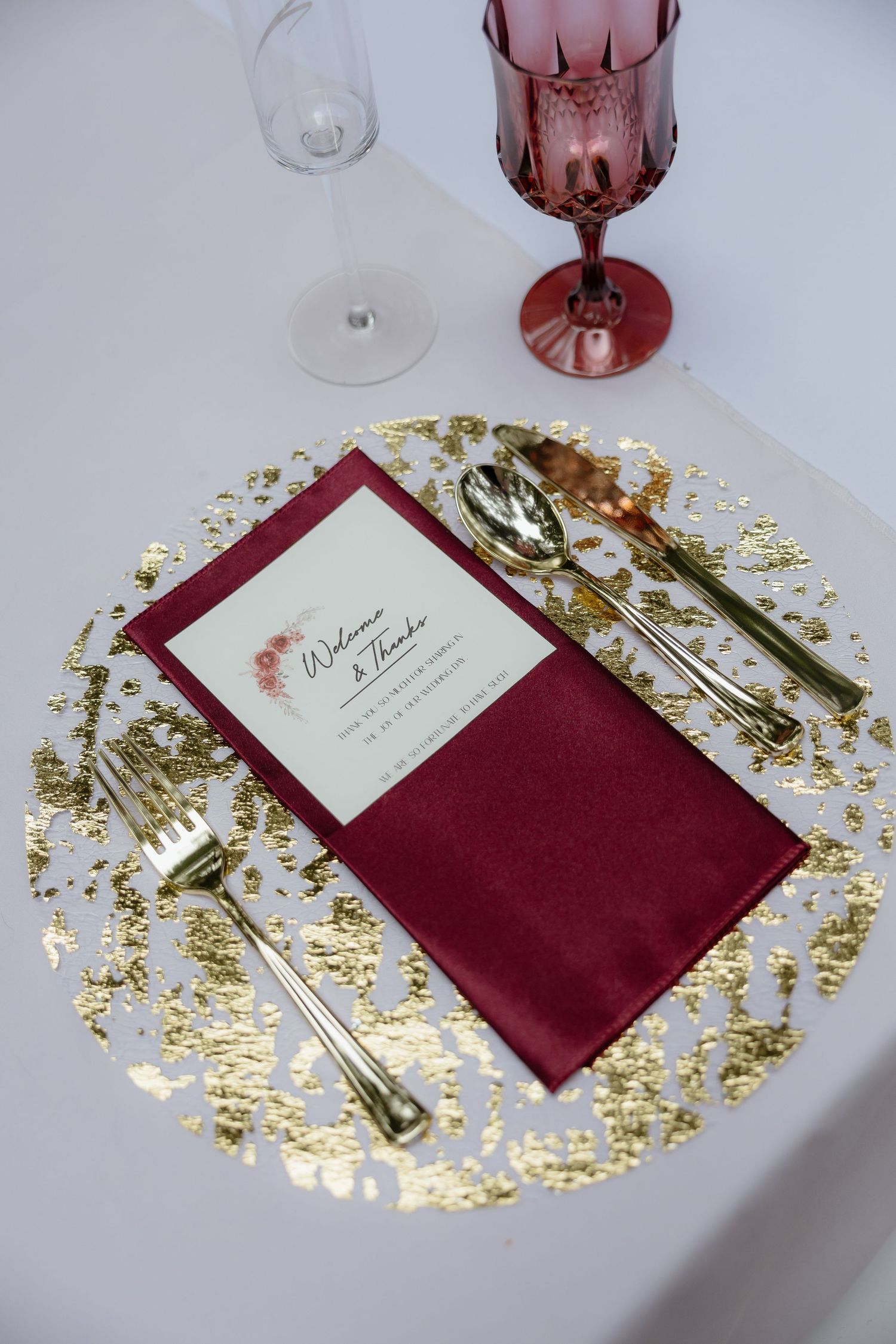Close-up of wedding place setting with gold leaf charger, burgundy napkin, gold utensils, and pink goblet on white table.