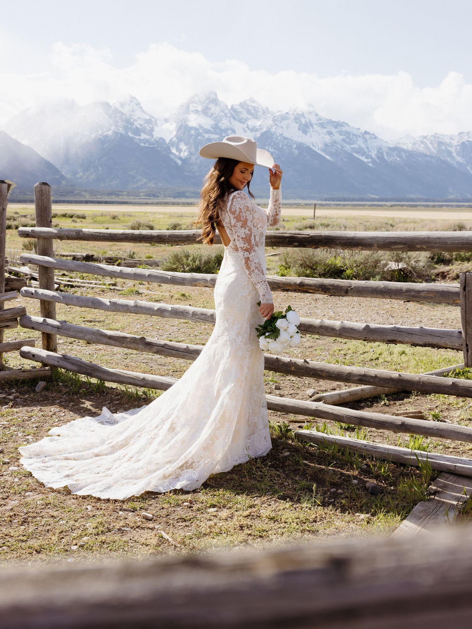 Bride in lace mermaid gown and white cowboy hat holds bouquet by wooden fence with snowy Teton peaks.