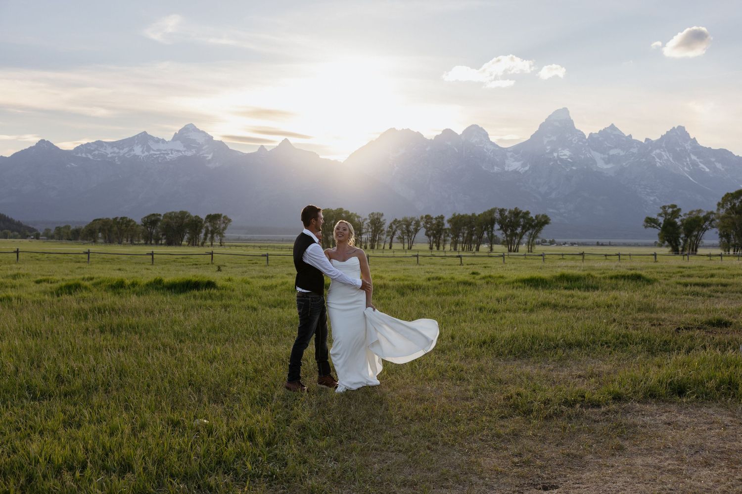 Bride and groom dance in green meadow at sunset with majestic Grand Teton mountains illuminated in background.