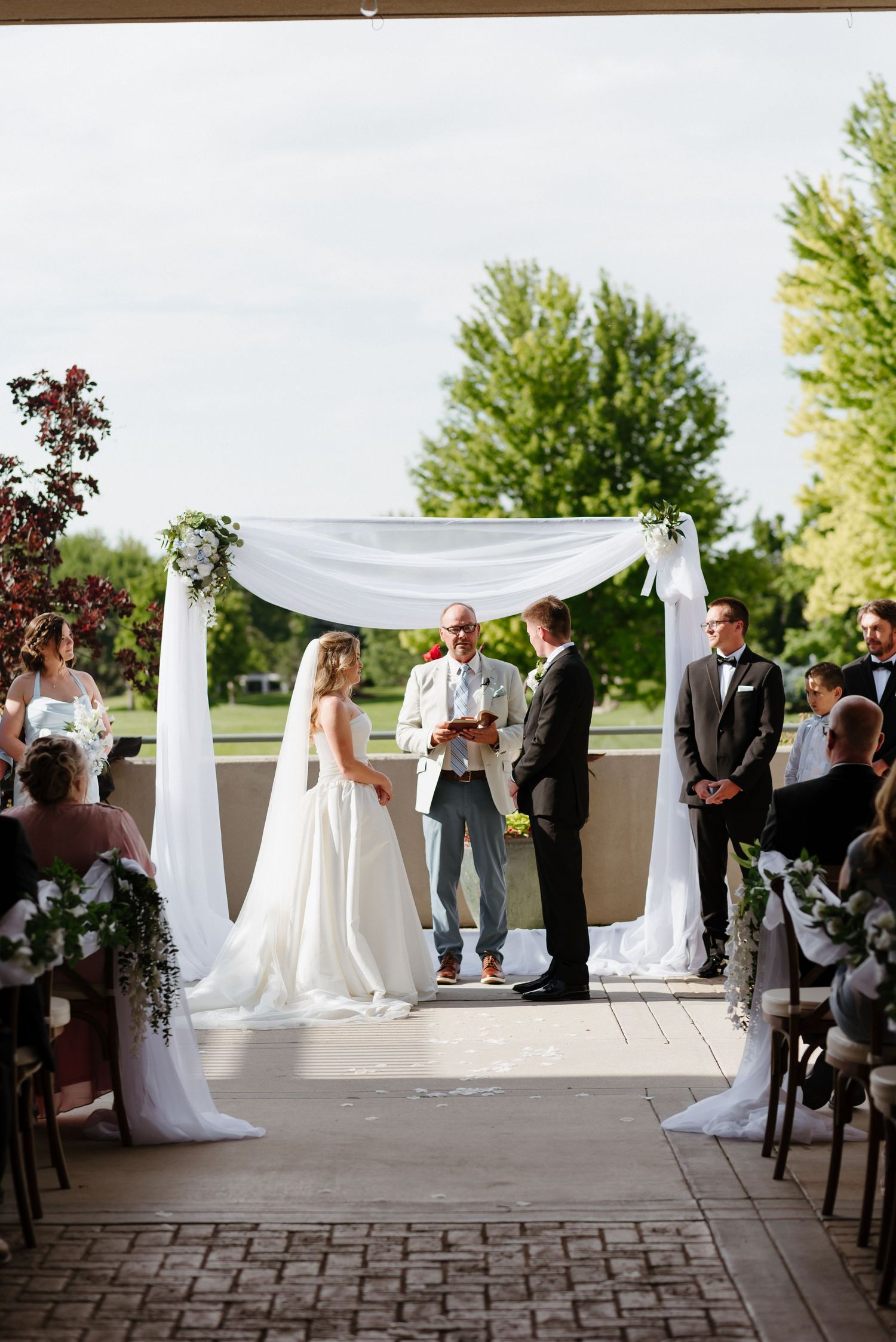 Bride and groom exchange vows under a white draped arch at an outdoor wedding ceremony.