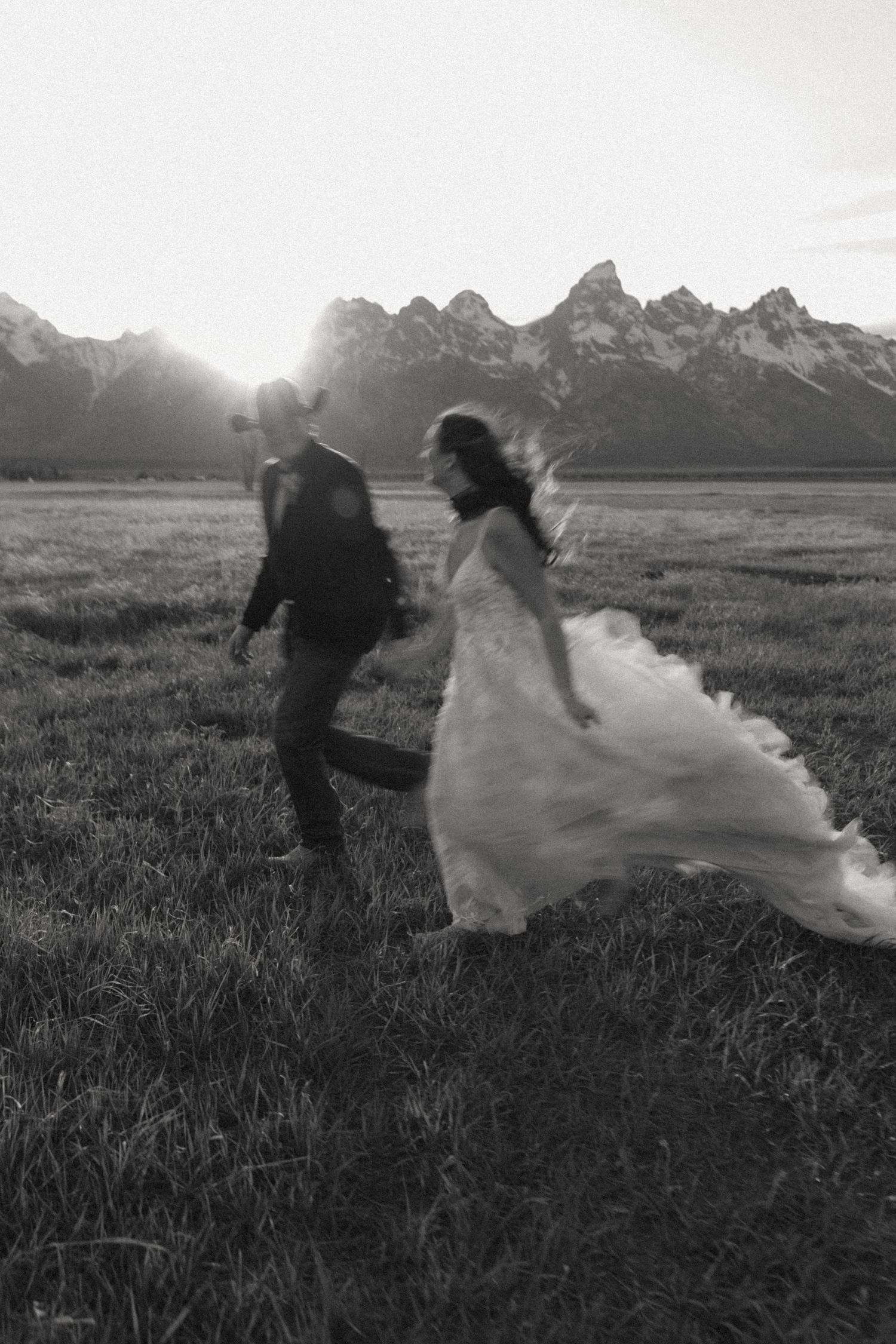 Black and white photo of bride and groom running through a field with the Grand Teton mountains at sunset.