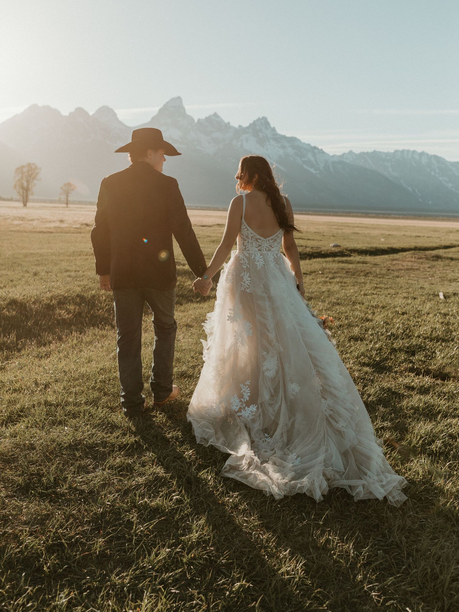 Bride and groom holding hands walking toward Grand Teton mountains in stunning backlit wedding photo.