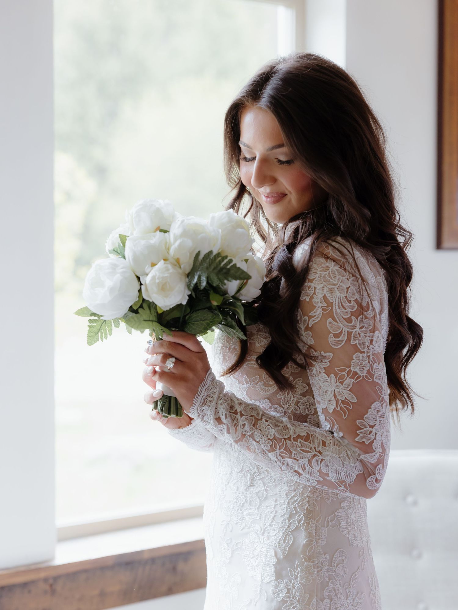 Bride in long-sleeve lace wedding gown smiling while holding a white peony bouquet by a bright window.
