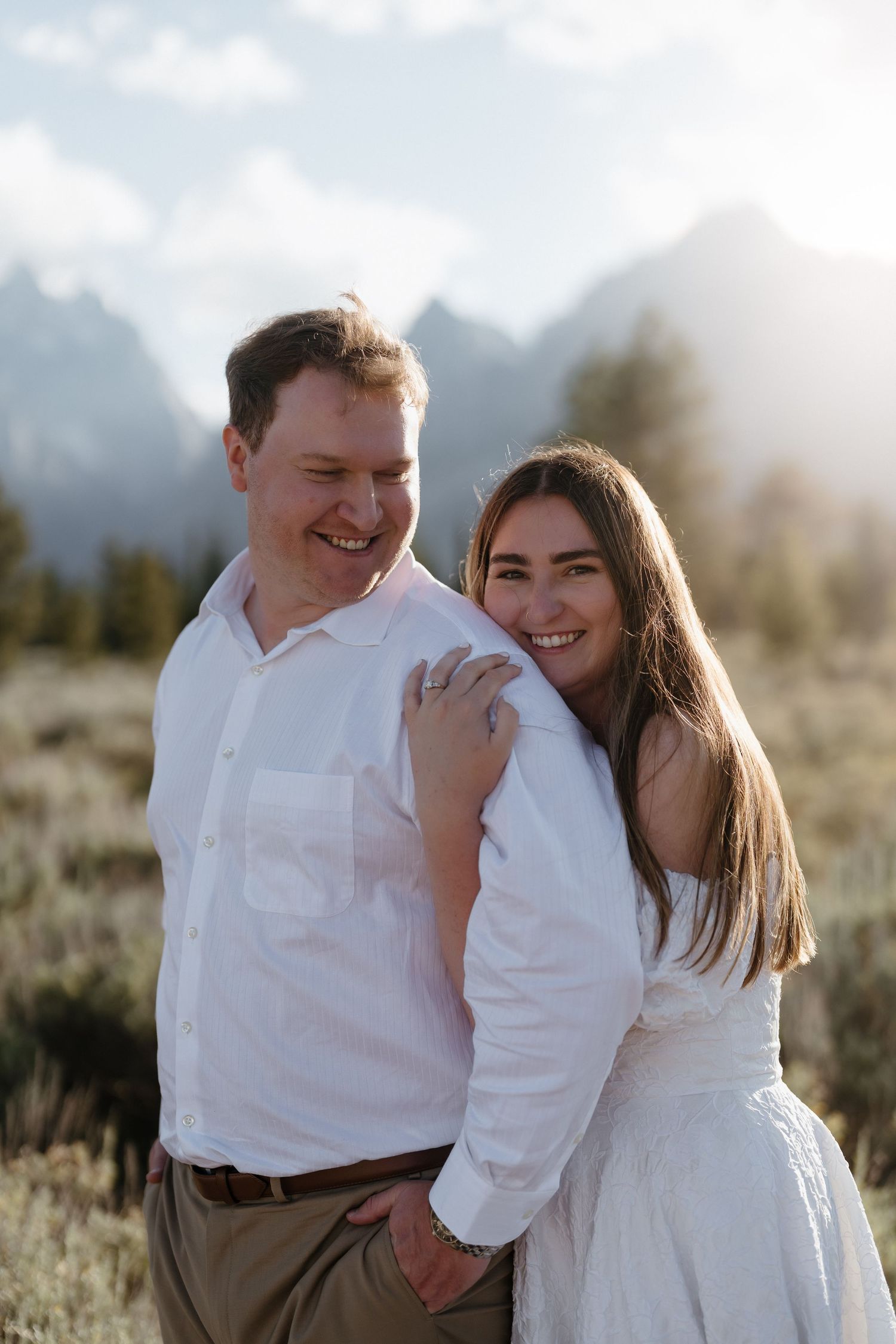 Smiling couple in white dress and shirt embrace outdoors with majestic mountain peaks glowing behind them.